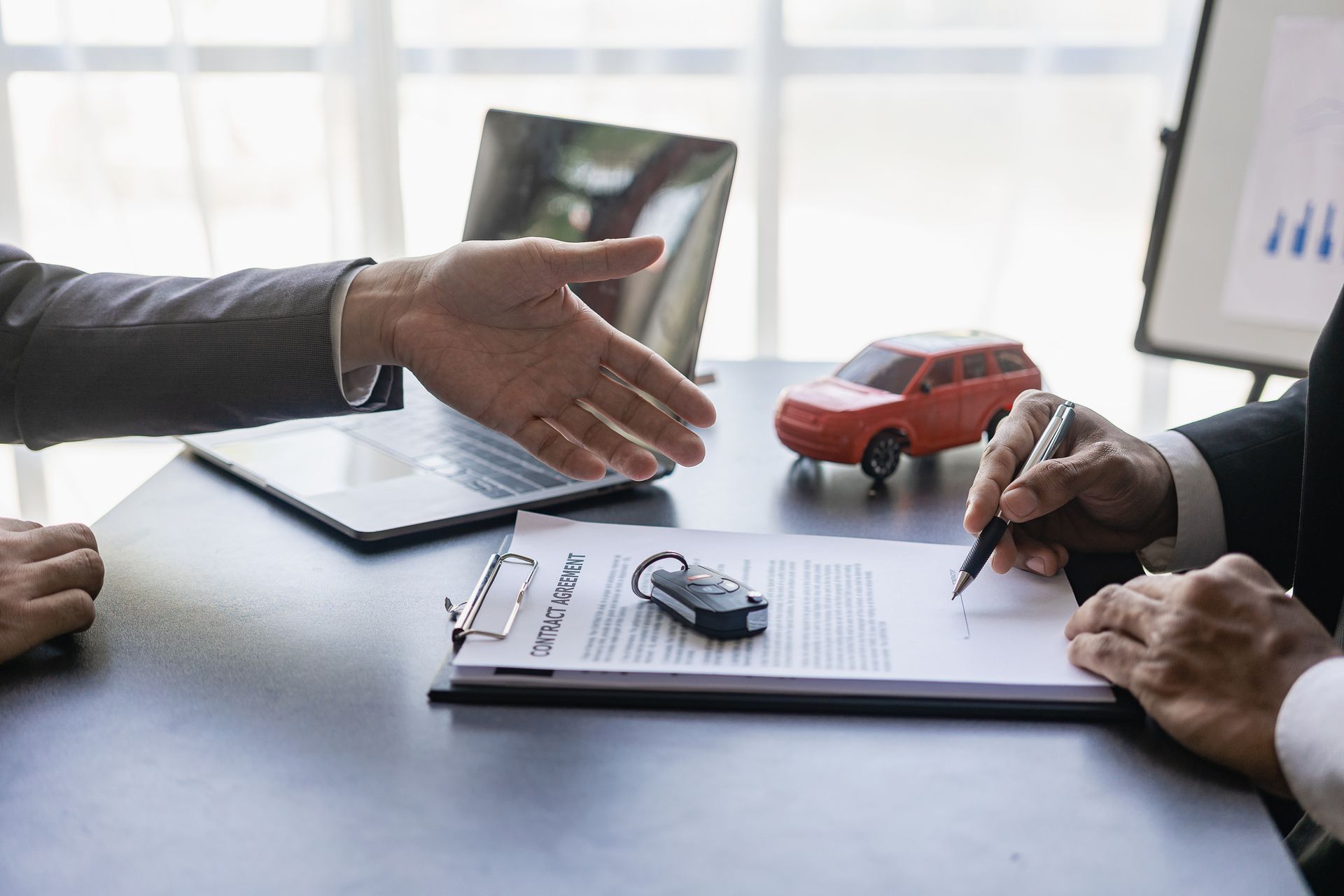 Hands discussing a car deal over documents with a toy red car and laptop on a desk