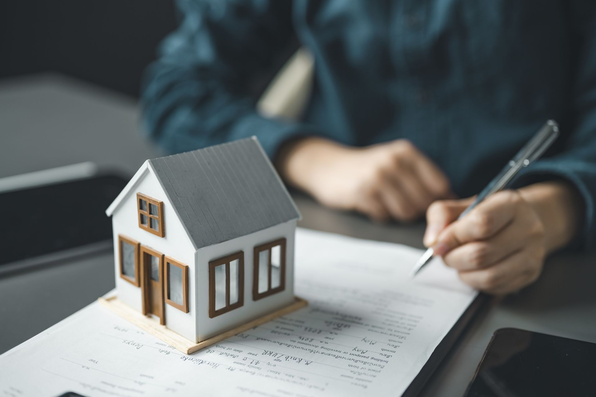 Miniature house on paperwork beside a person writing with a pen at a desk