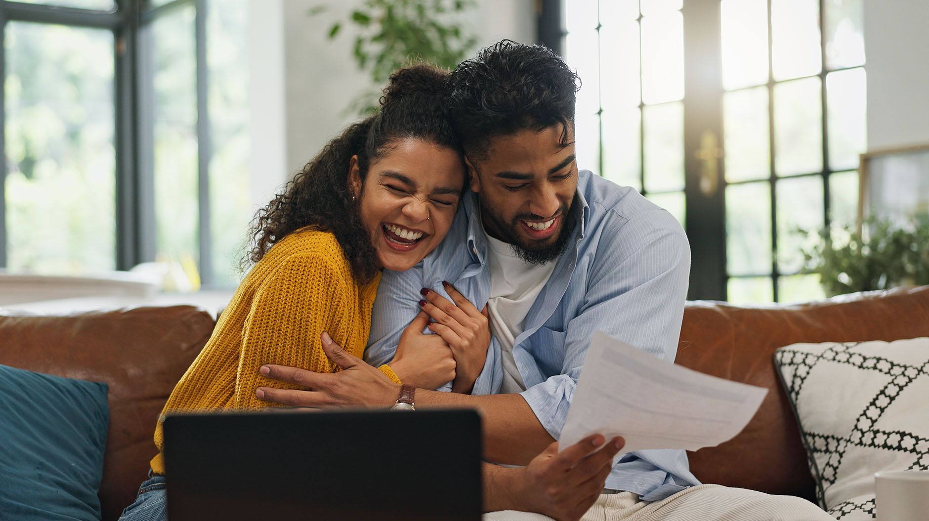 Couple laughing while looking at papers, sitting on a couch near a laptop.