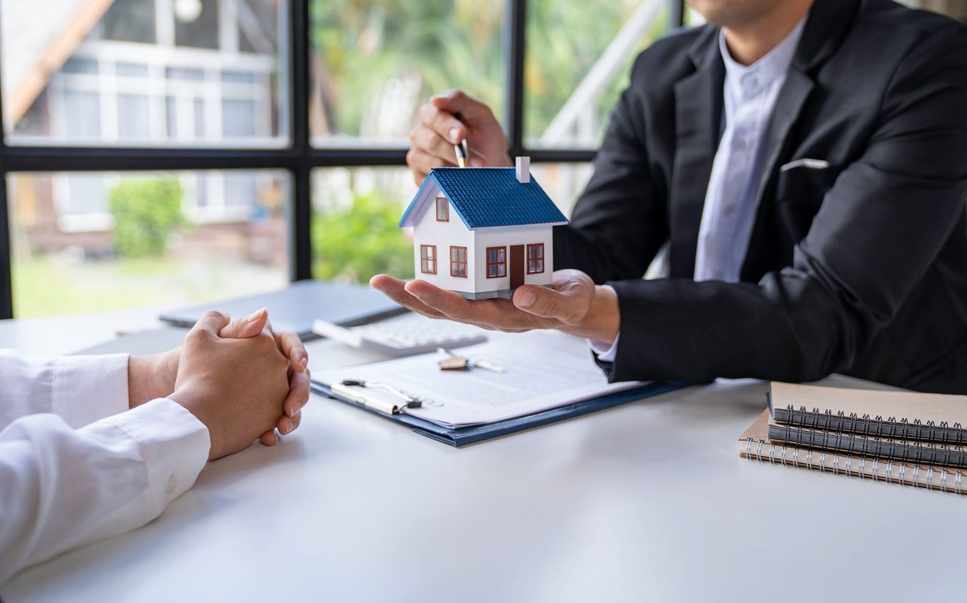 Real estate agent holding house model, presenting to a client at a desk.