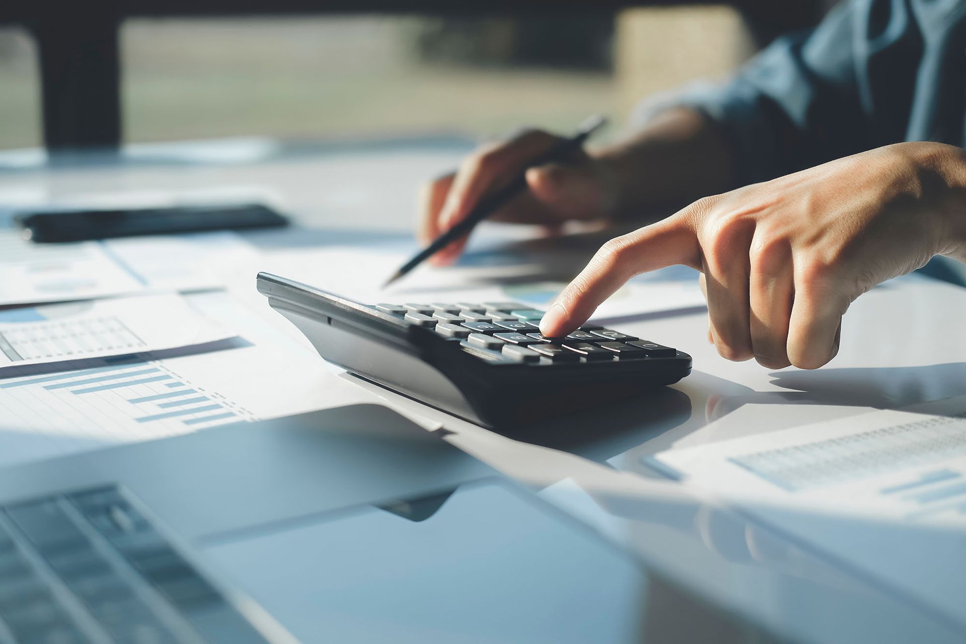 Person calculating with a calculator, documents and a laptop in a bright office.