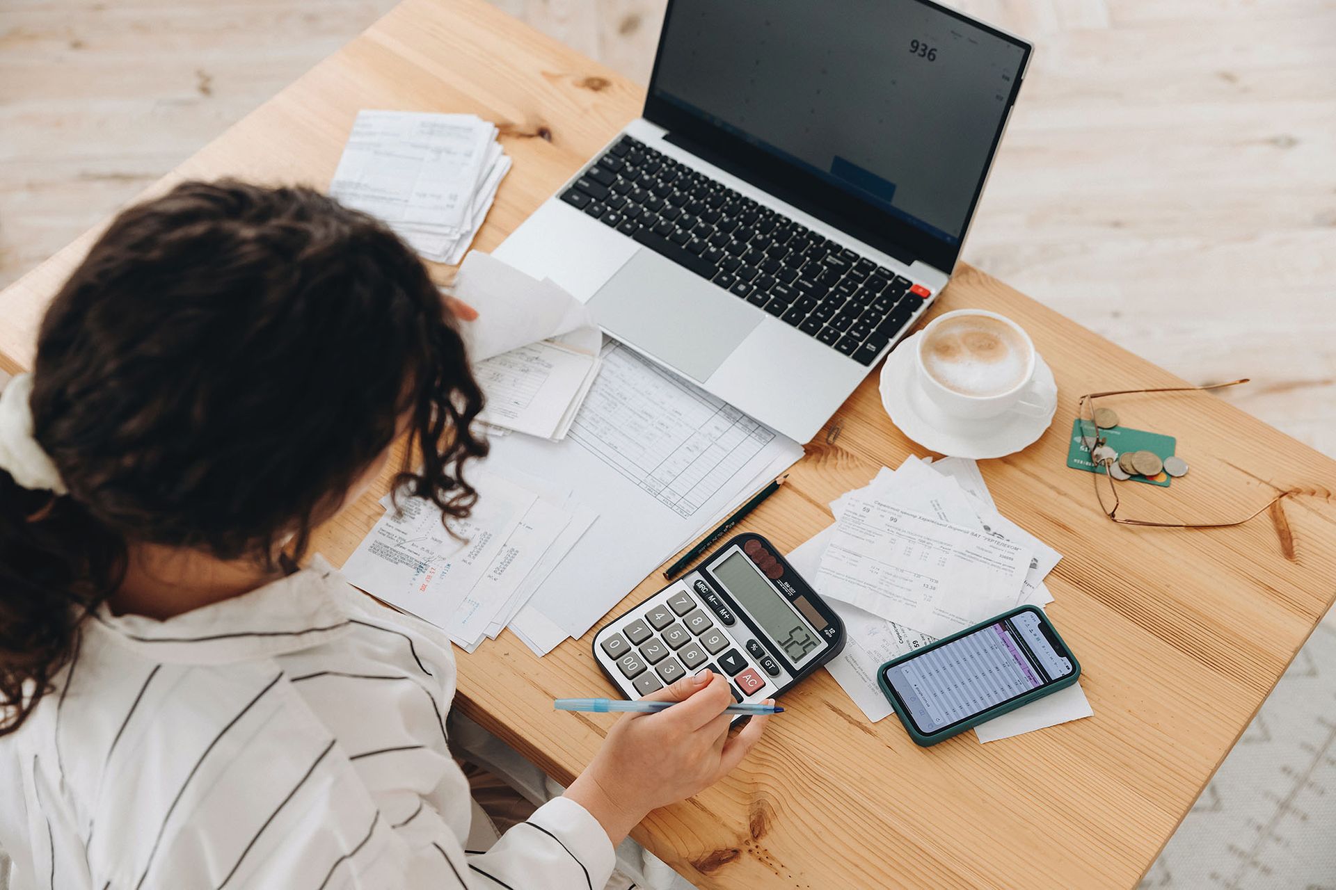 Woman working on laptop and using calculator, surrounded by papers, phone, and coffee.