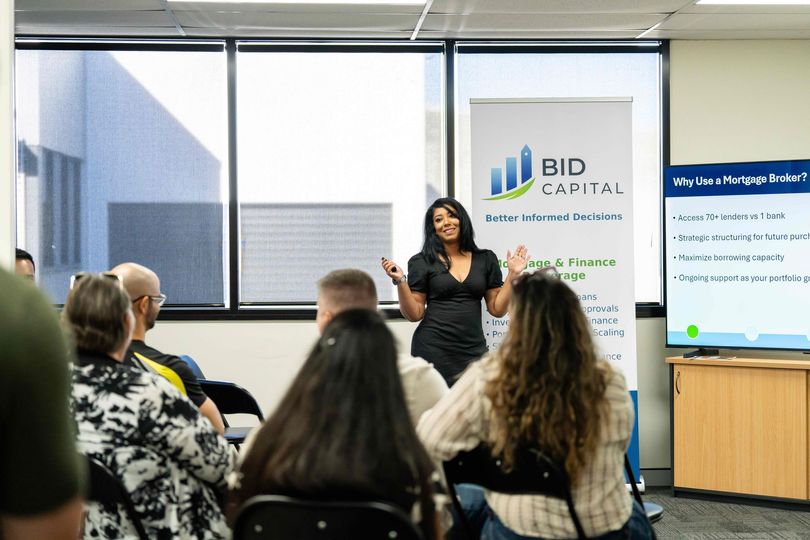Presenter speaking to seated audience in a bright office meeting room with BID Capital banner and presentation screen