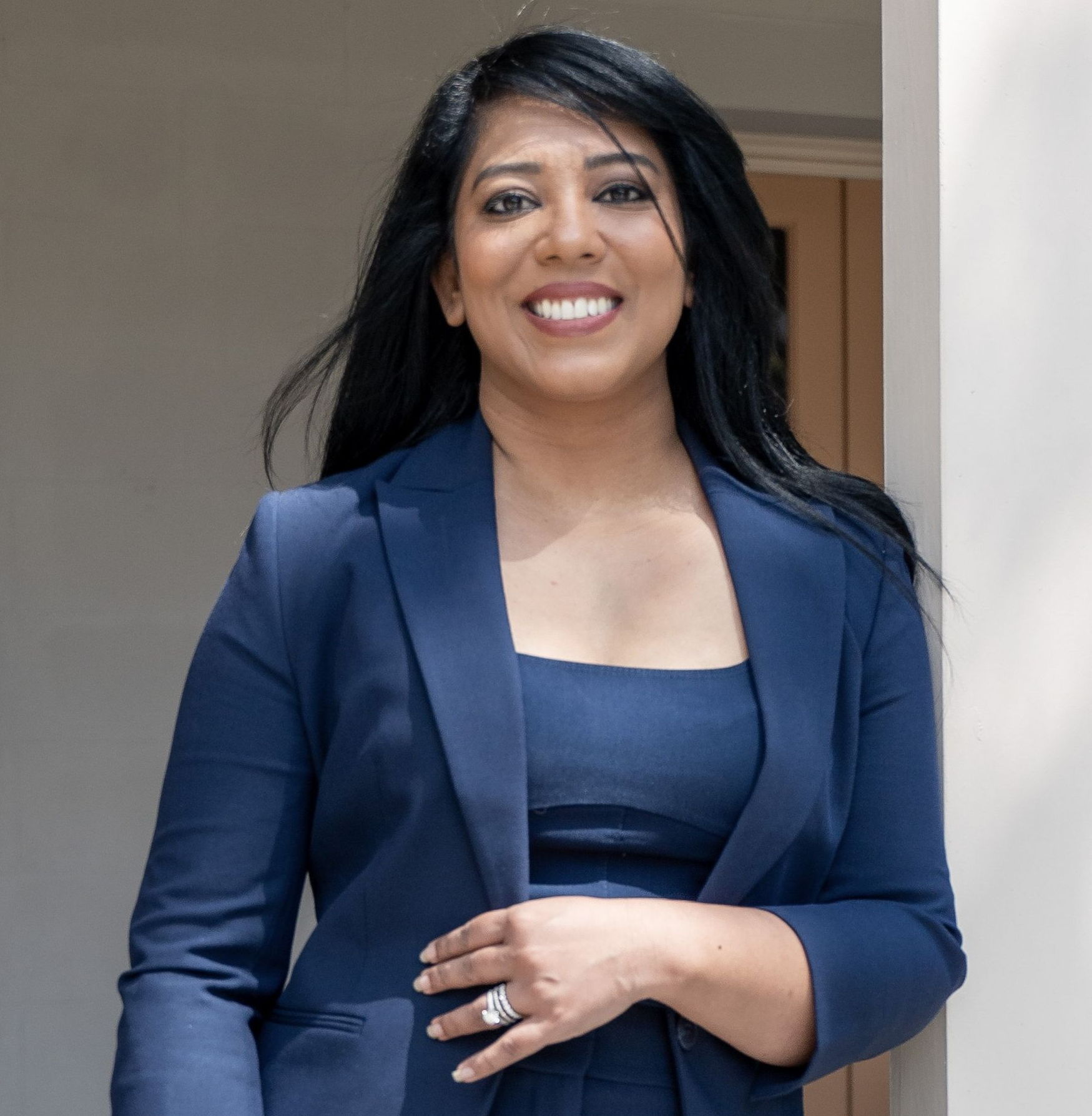Woman in blue blazer and top smiles, leaning against a white pillar outside.