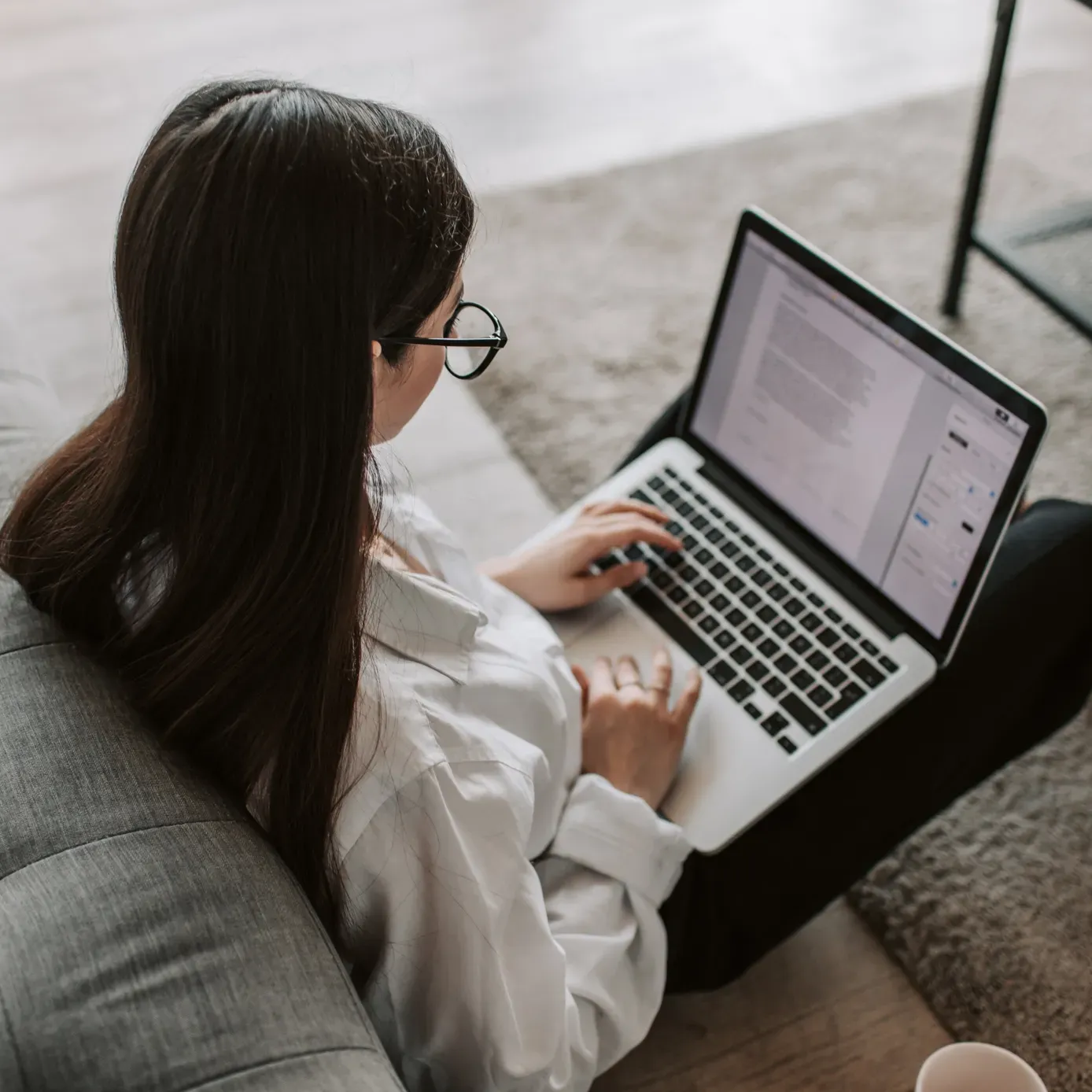 Woman sitting on a couch, using a laptop, typing. Wearing glasses and a white shirt.