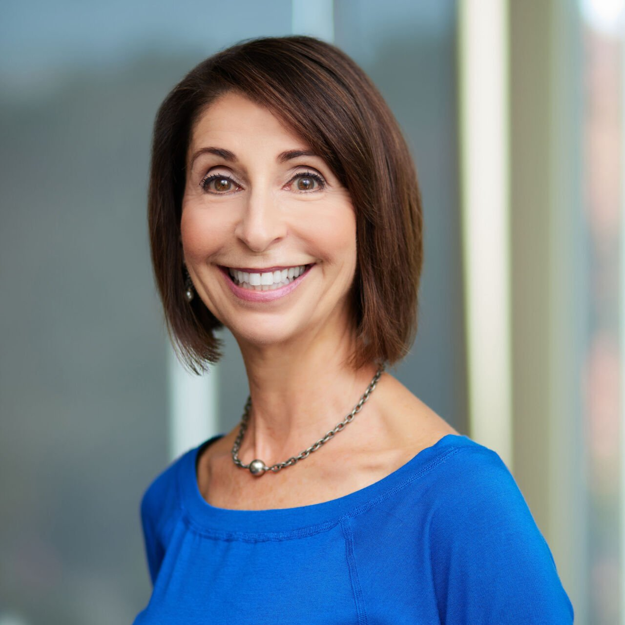 Woman with brown hair, smiling in a blue shirt, wearing a silver necklace.