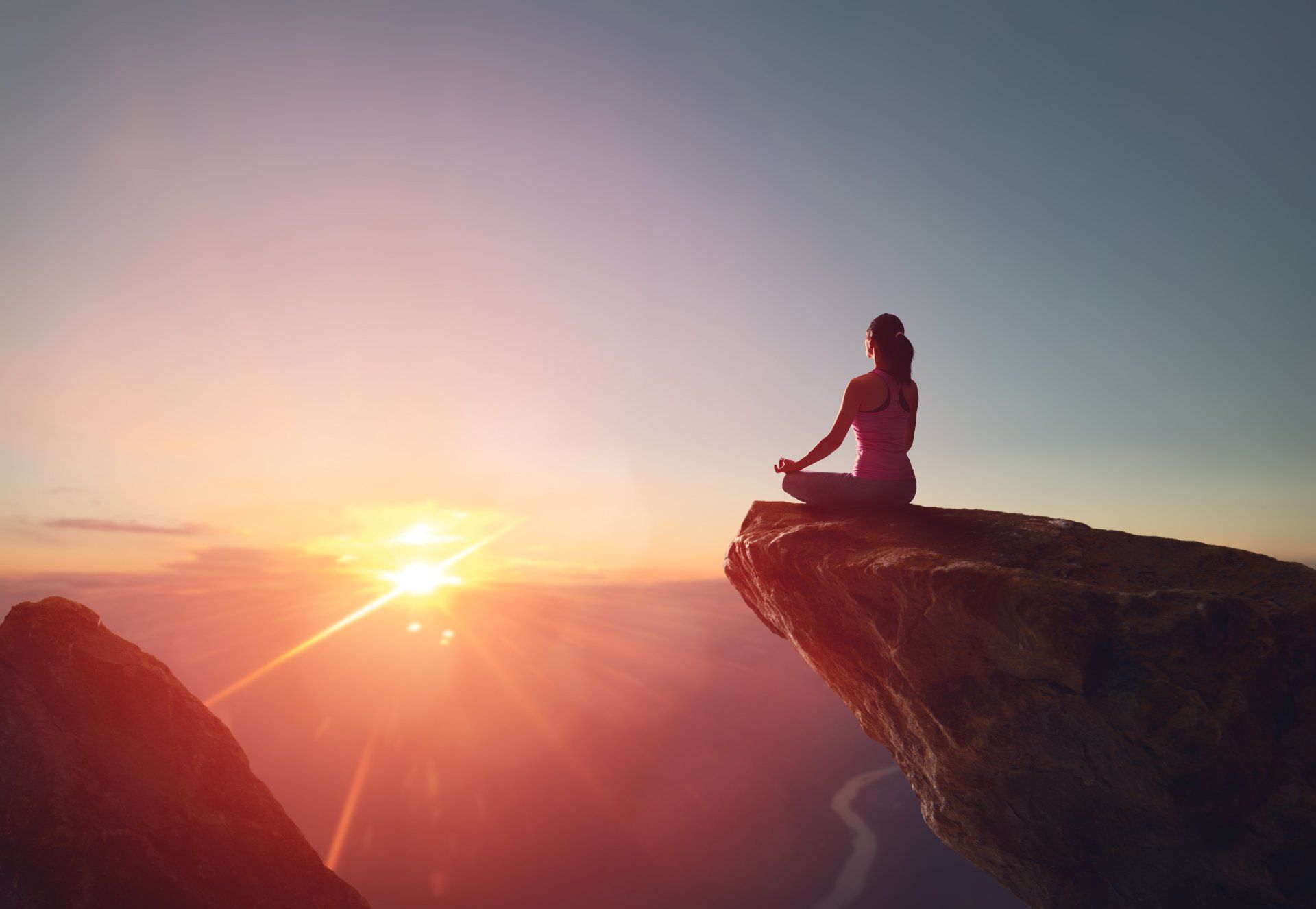 Woman meditates on cliff edge at sunset; orange sky, mountain backdrop.