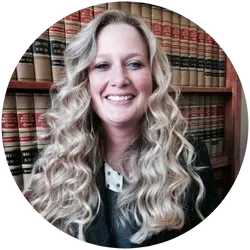 A woman with long blonde hair is smiling in front of a bookshelf filled with books.