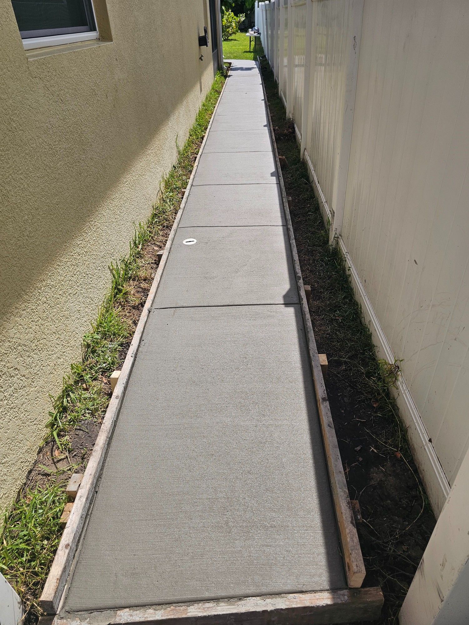 Concrete walkway between a tan building and a white fence with green grass and weeds along the sides.