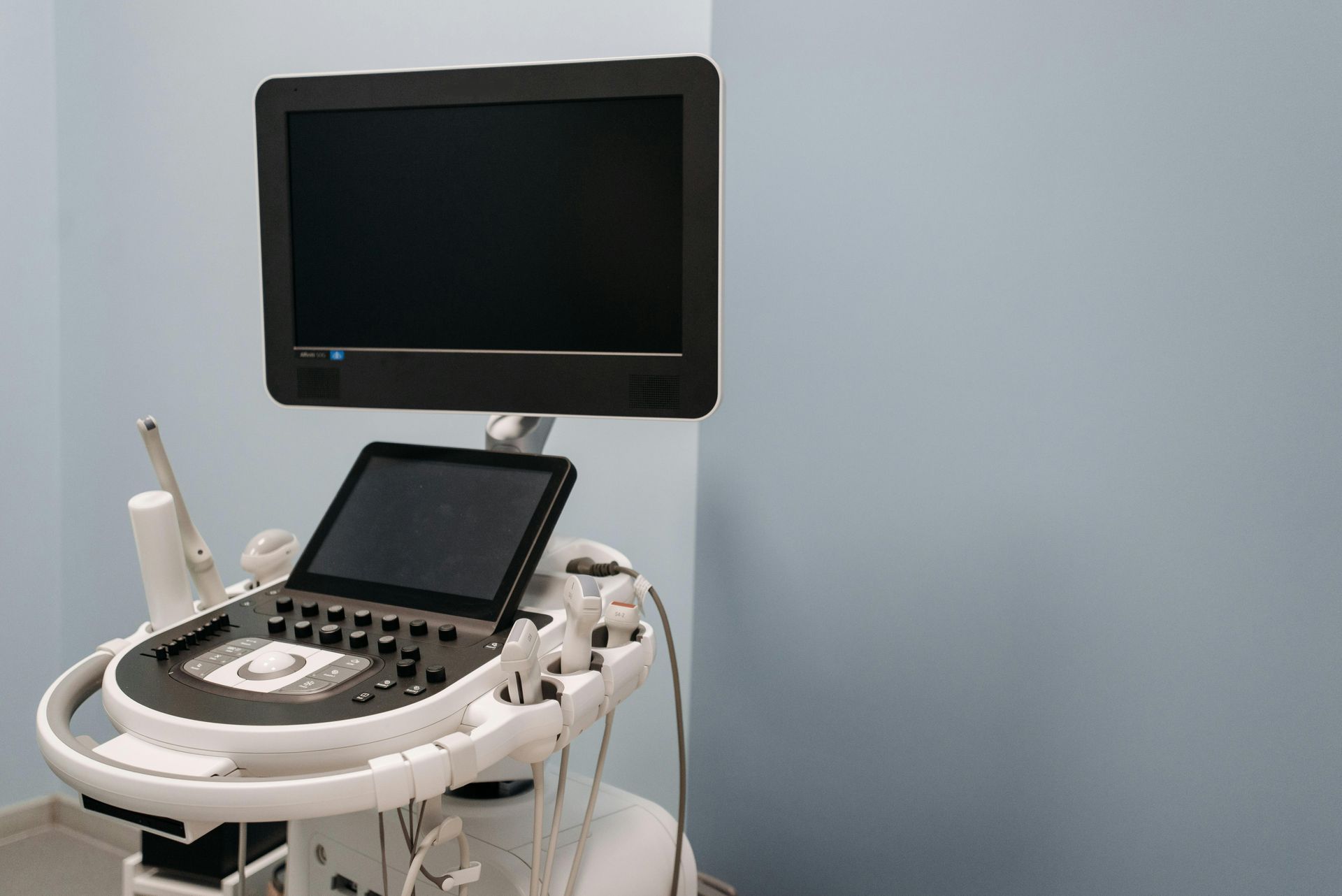 An ultrasound machine with a large screen and control console stands in a room with light blue walls.