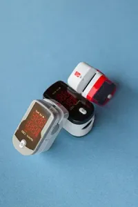 Three finger-clip pulse oximeters in gray, black, and red/white, arranged diagonally on a plain blue background.