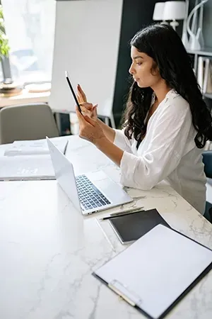 A person with long dark hair sits at a desk working on a laptop while holding a stylus and a tablet.