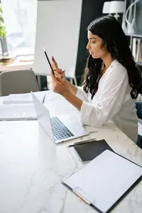 A person with long dark hair sits at a desk working on a laptop while holding a stylus and a tablet.