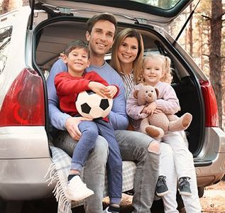Family sitting in the open trunk of a car, smiling and holding a soccer ball and teddy bear.