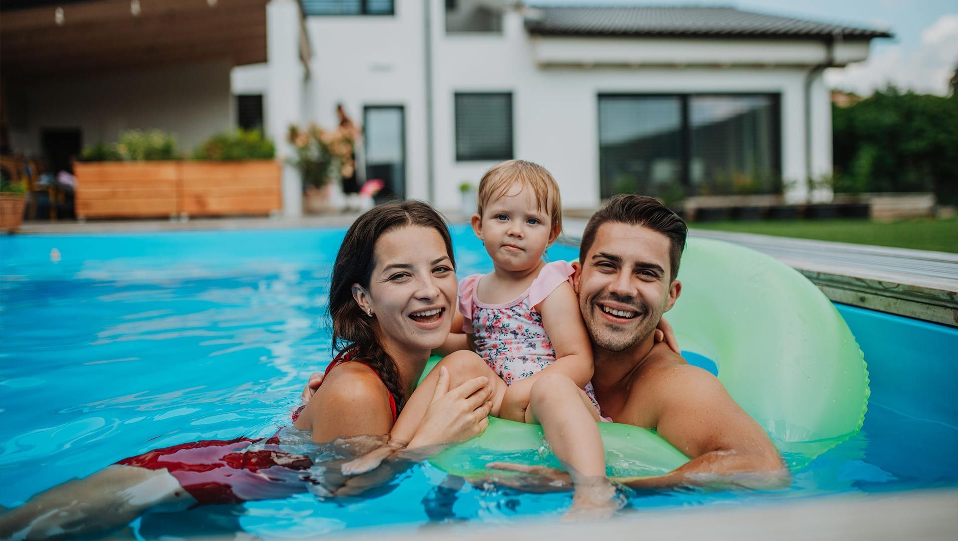 Family smiling in a backyard pool with a green float and a house in the background