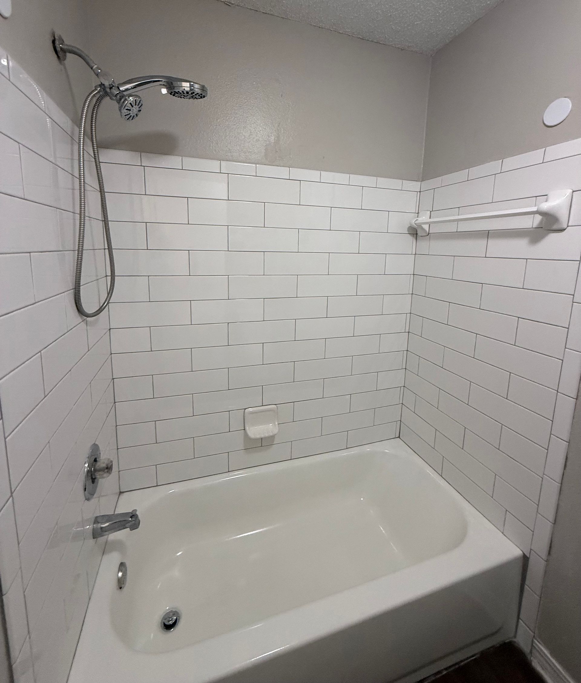 White tiled bathtub and shower stall with chrome showerhead and grab bar in a small bathroom