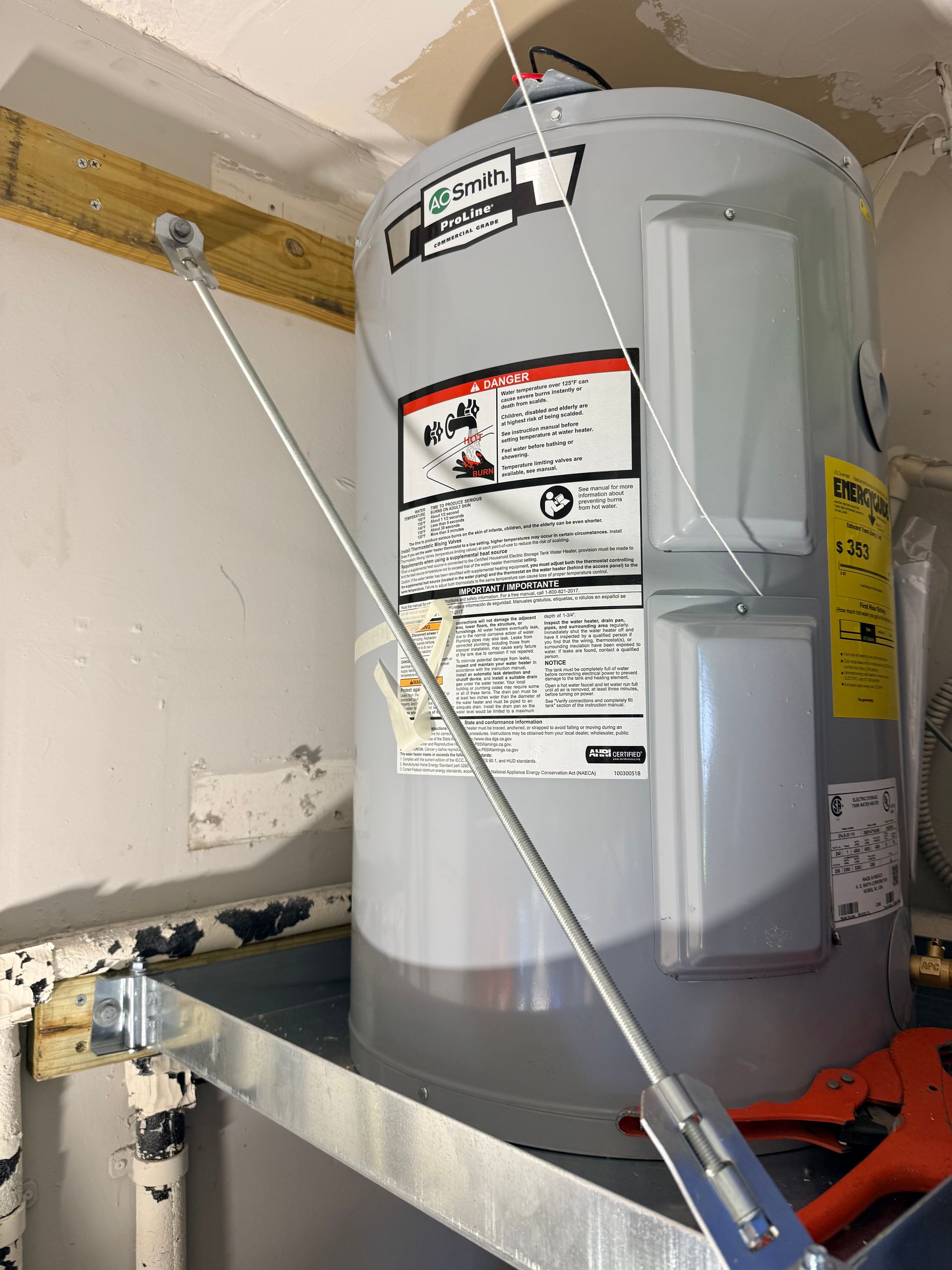 Water heater installed on a metal platform in a utility room, with a support strap and labels visible.
