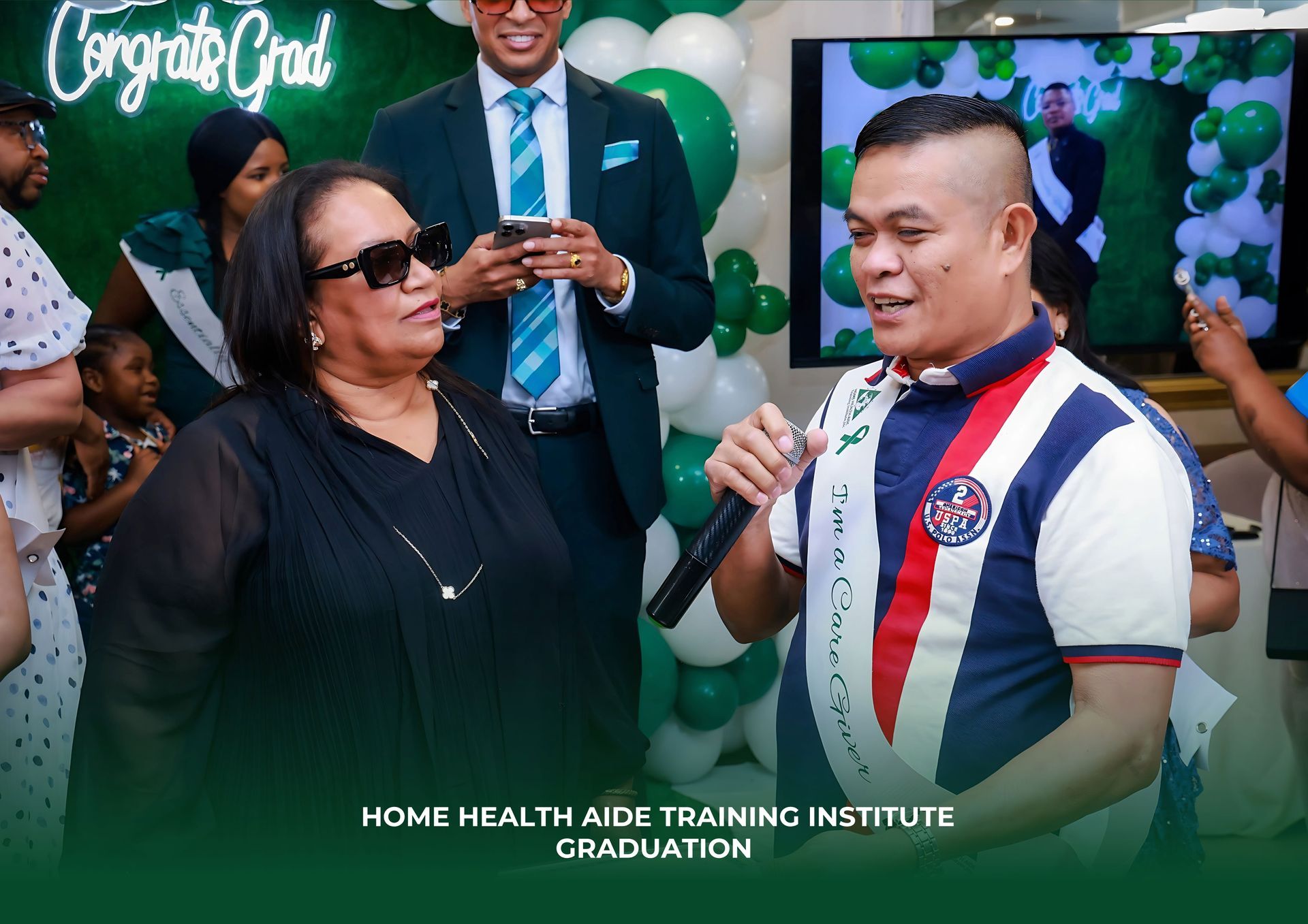 A graduate speaks into a microphone while a woman in sunglasses listens at a Home Health Aide Training Institute event.