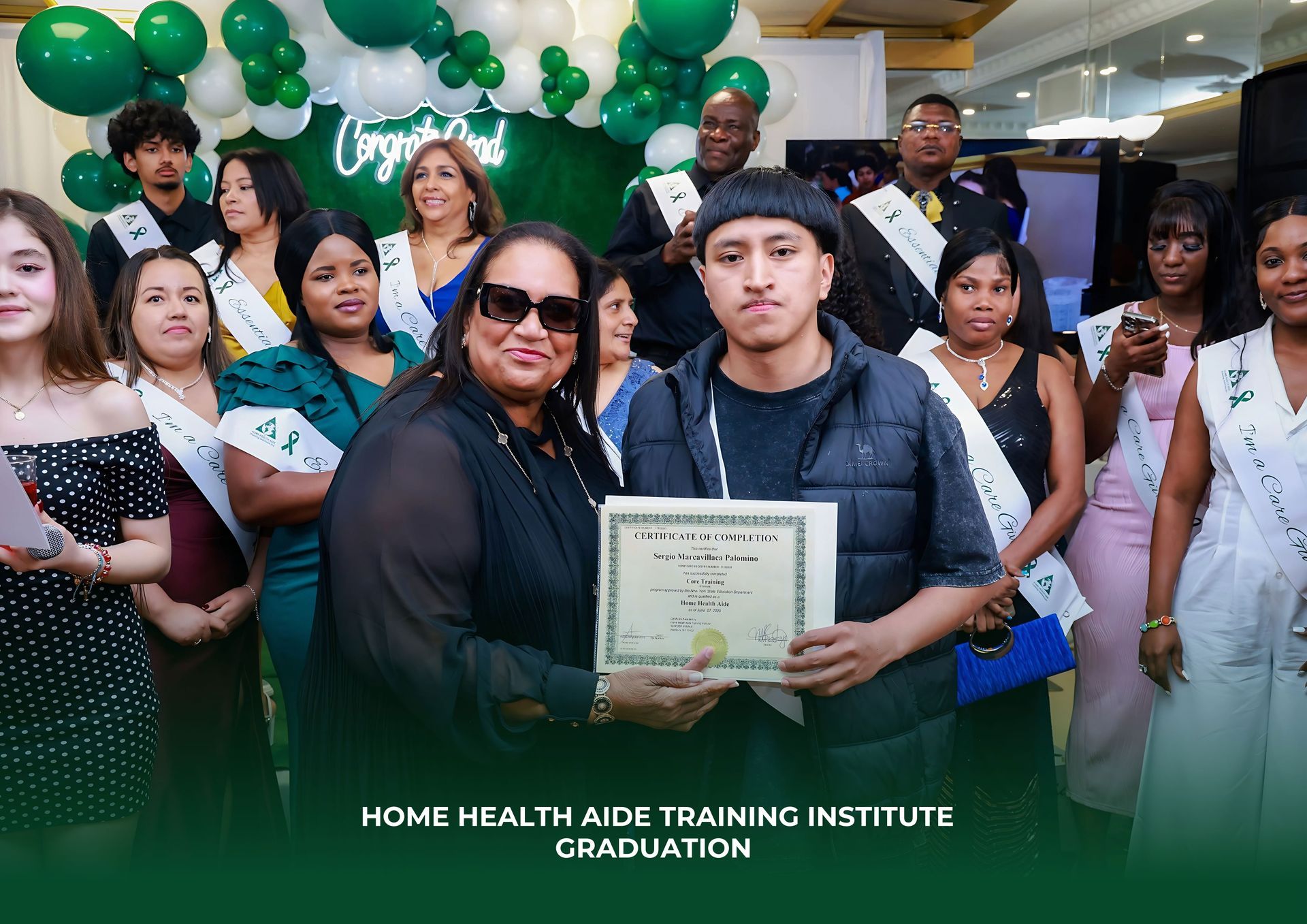 Graduates at a ceremony, with one student holding a certificate while an instructor looks on against a balloon backdrop.