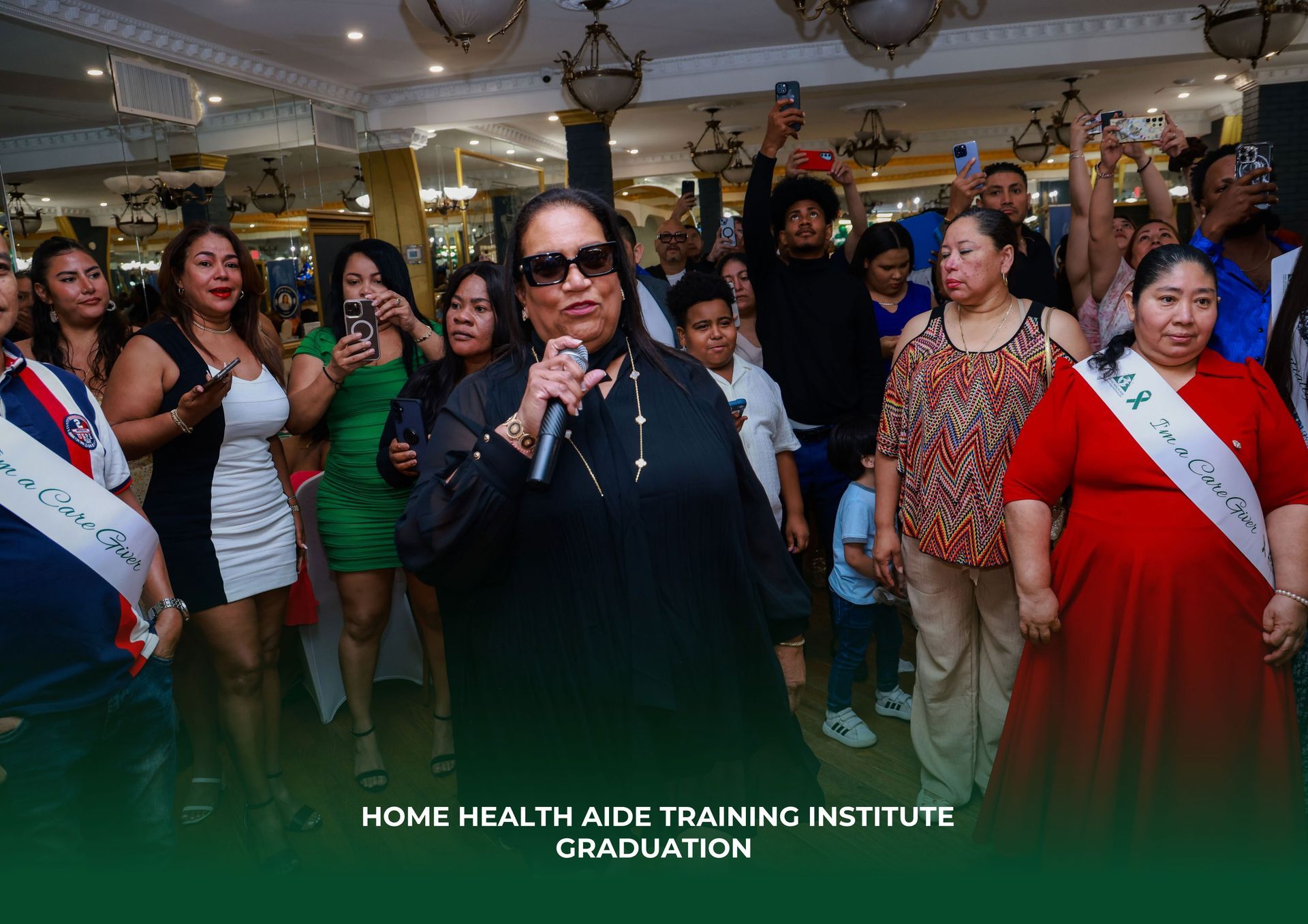 A group of graduates in sashes stand at a Home Health Aide Training Institute graduation ceremony while one person speaks.