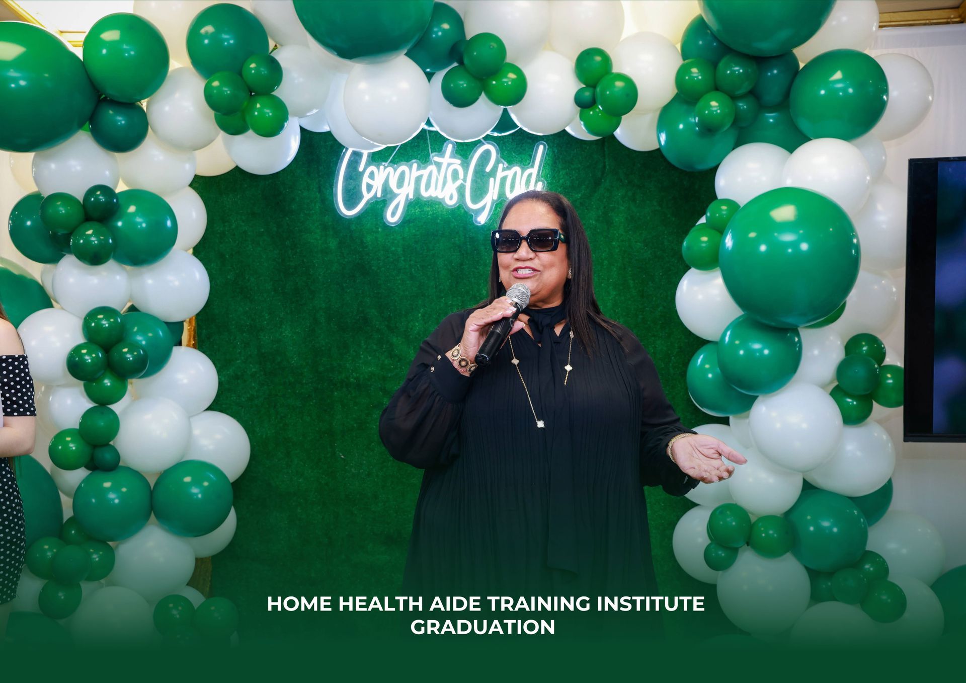 A person in black clothing and sunglasses speaks into a microphone before a green grass backdrop with a graduation sign.