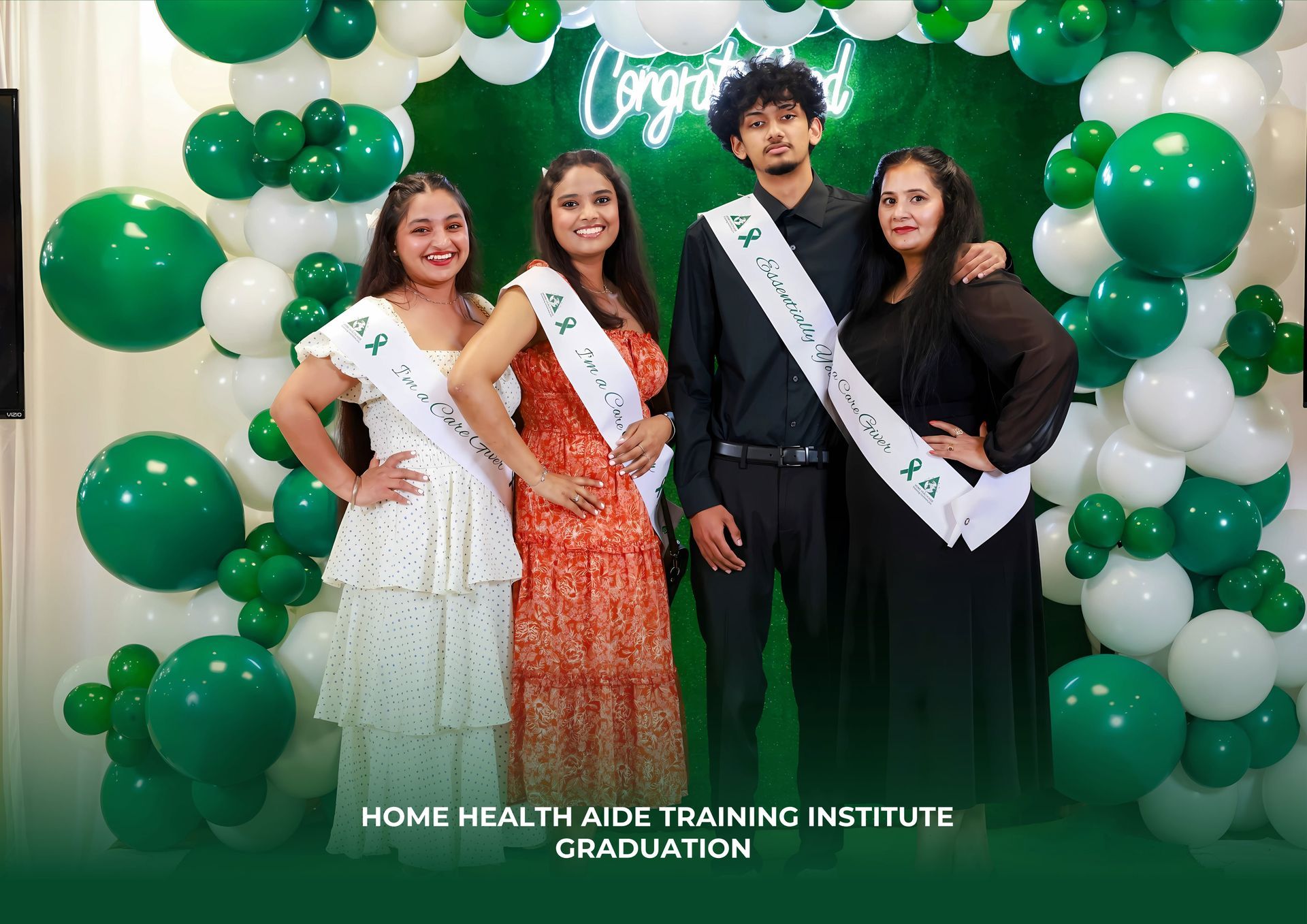 Four graduates in sashes stand before a green wall and balloon arch at a Home Health Aide Training Institute graduation.