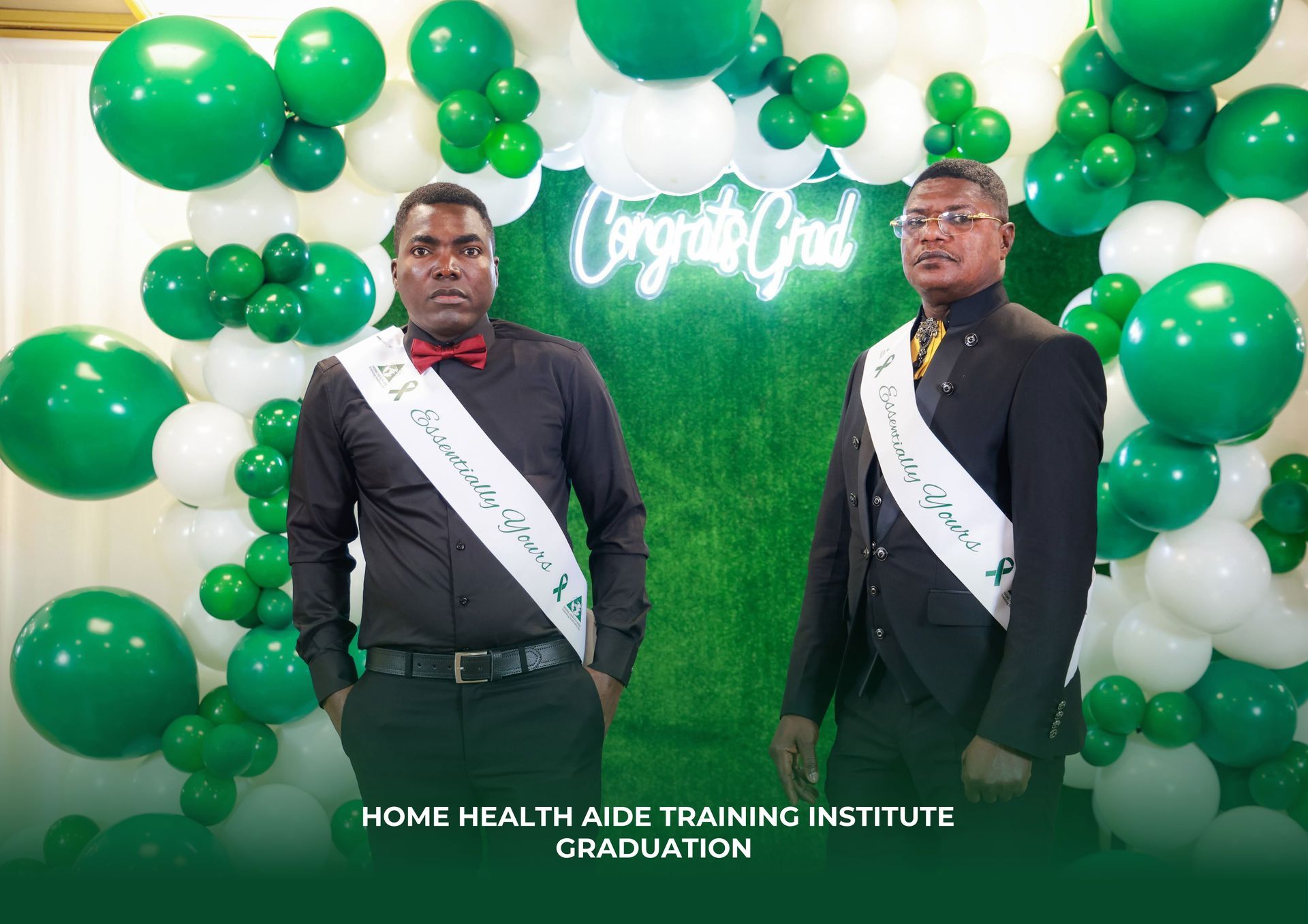 Two graduates wear sashes in front of a green balloon arch and a neon 