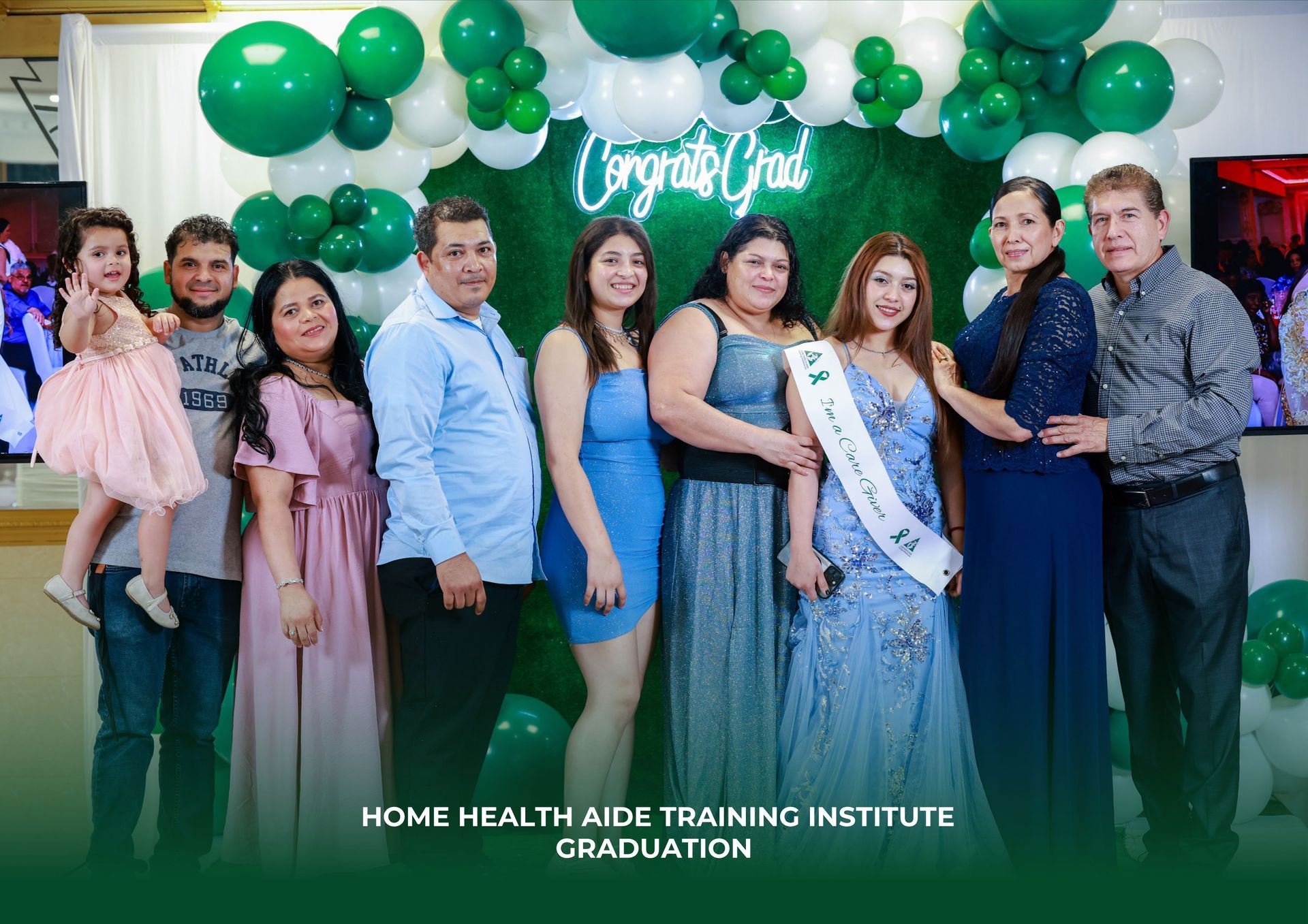 A family celebrates a graduate wearing a sash at a Home Health Aide Training Institute ceremony with green balloons.
