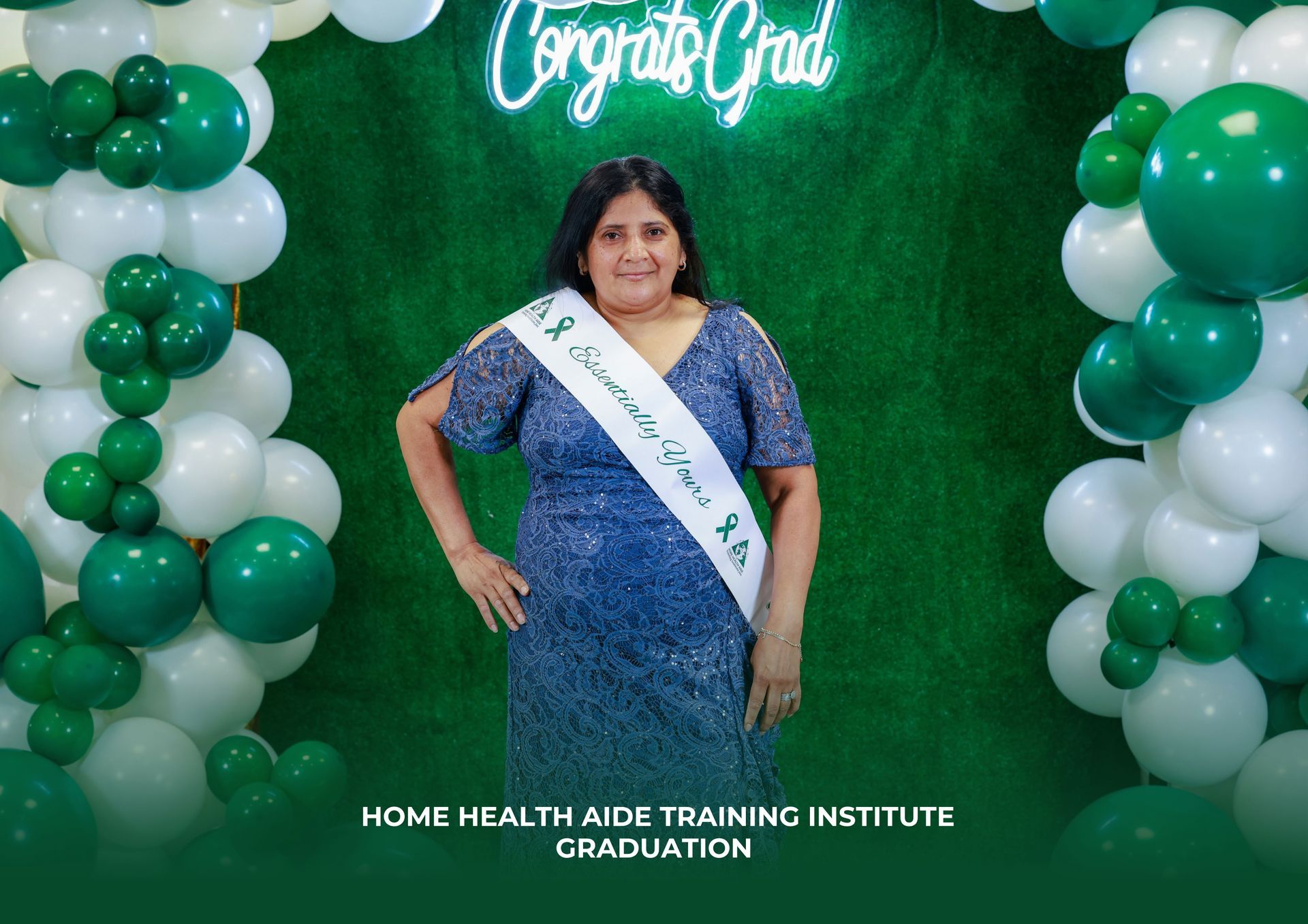 Graduate wearing a sash and blue dress stands before a green wall with balloons and a 