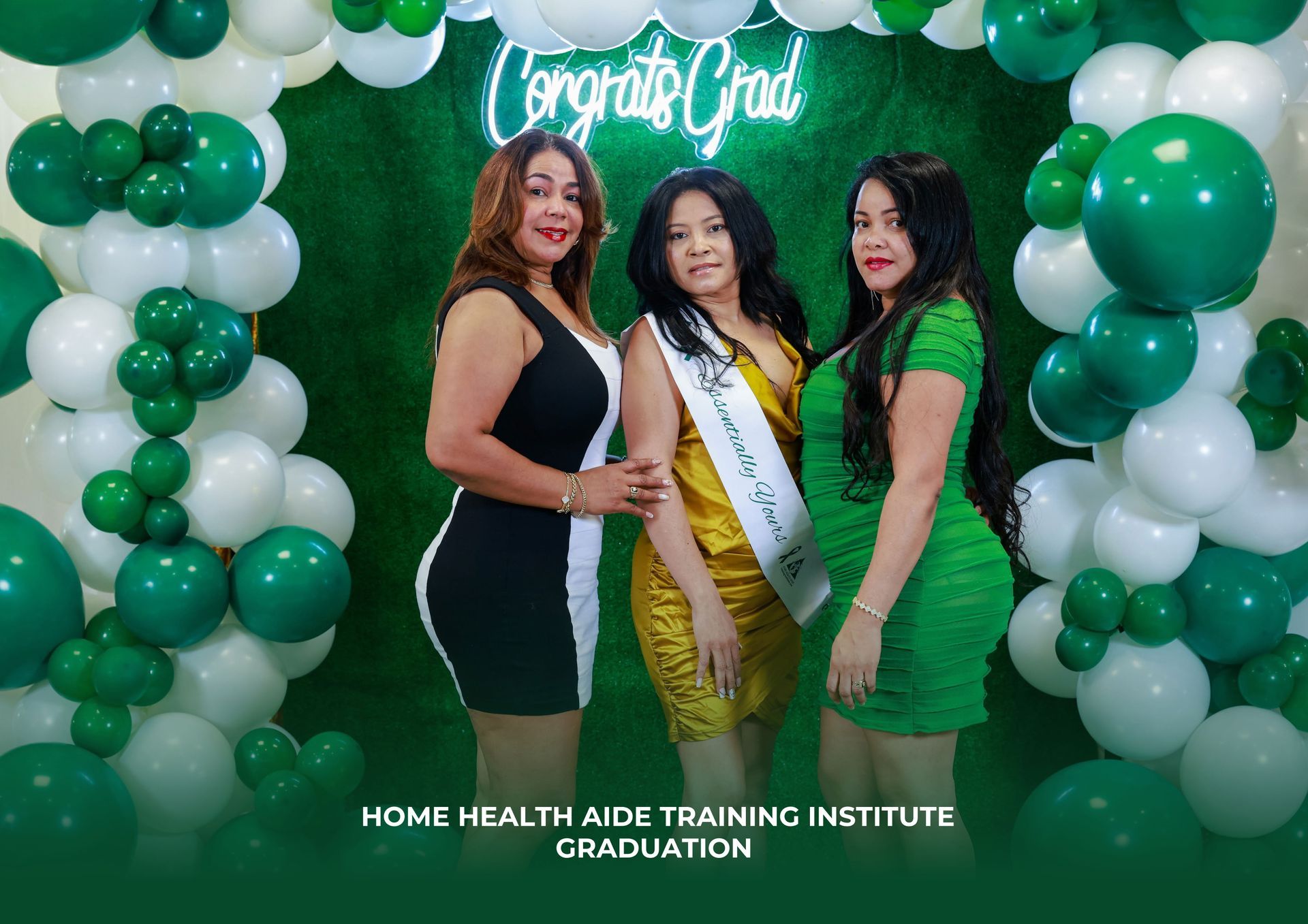Three people pose for a graduation photo in front of a green balloon arch and a neon 