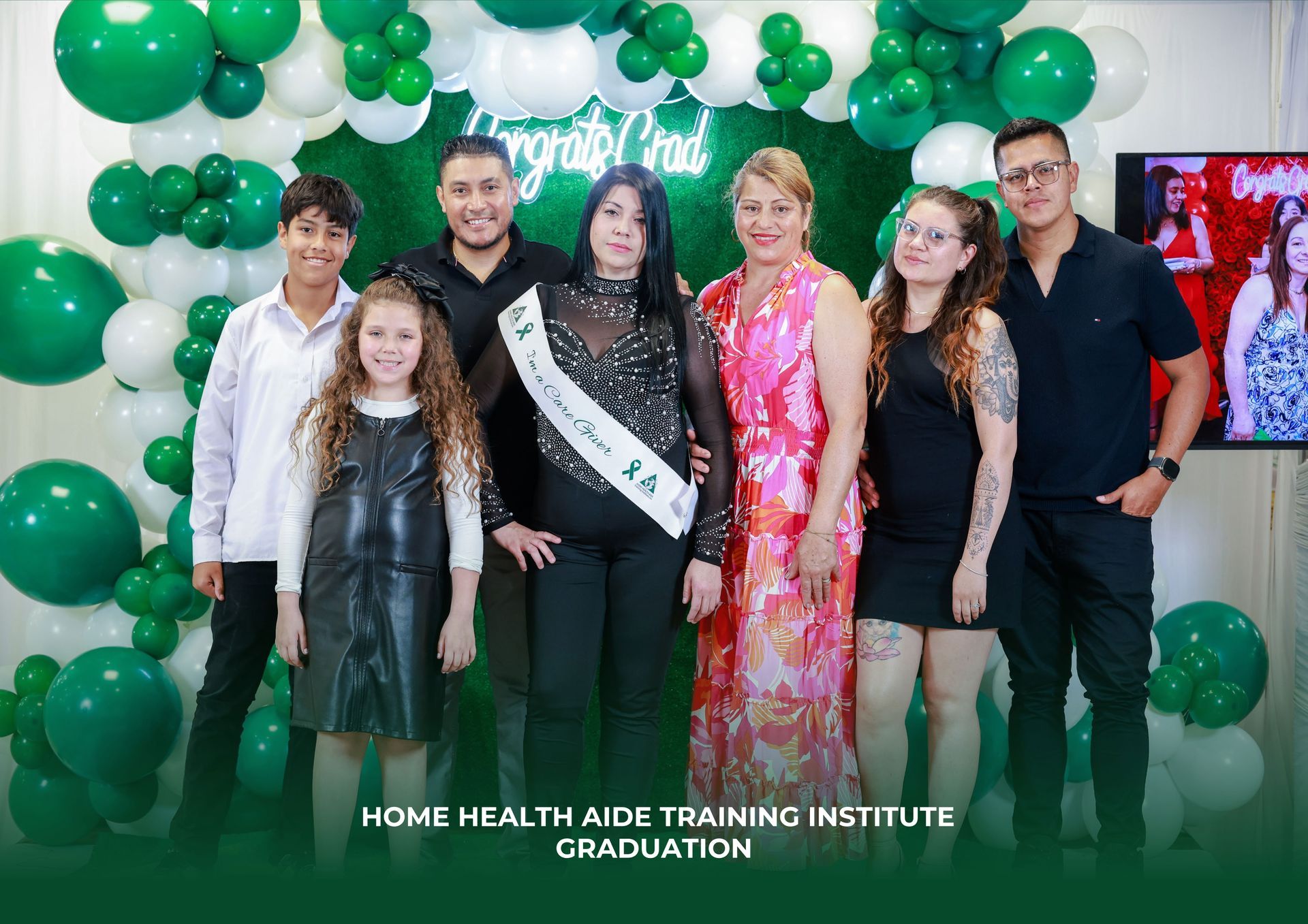 A family poses for a photo at a Home Health Aide Training Institute graduation, surrounded by green and white balloons.