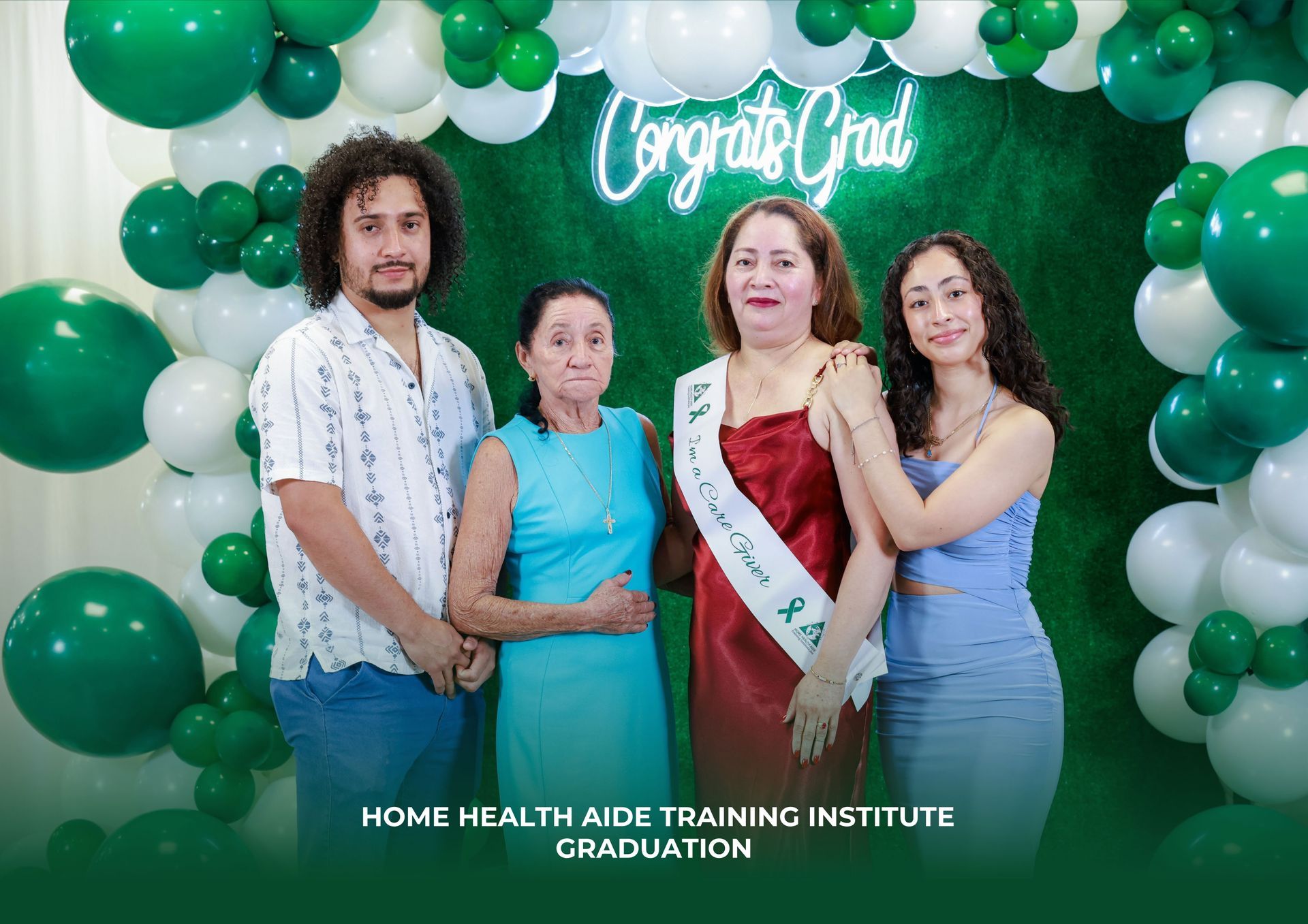 Four people pose for a graduation photo against a green balloon backdrop with a 