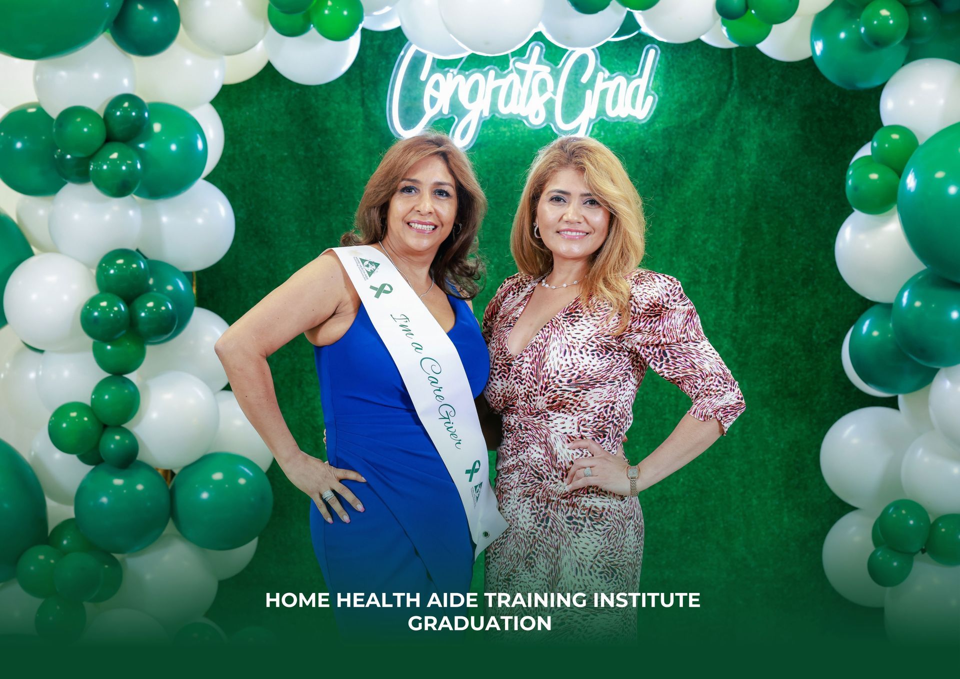Two women smiling in front of a green balloon arch with a 