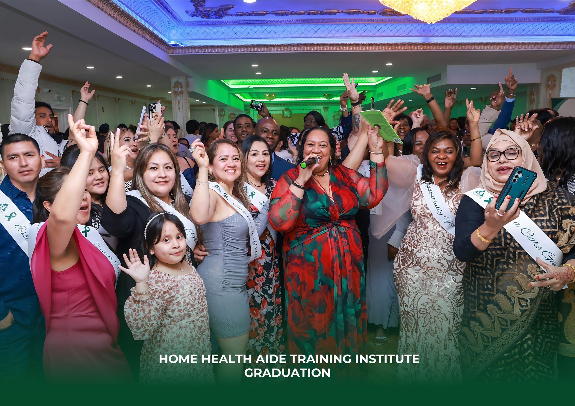 A group of graduates at the Home Health Aide Training Institute ceremony, smiling and cheering in an event hall.