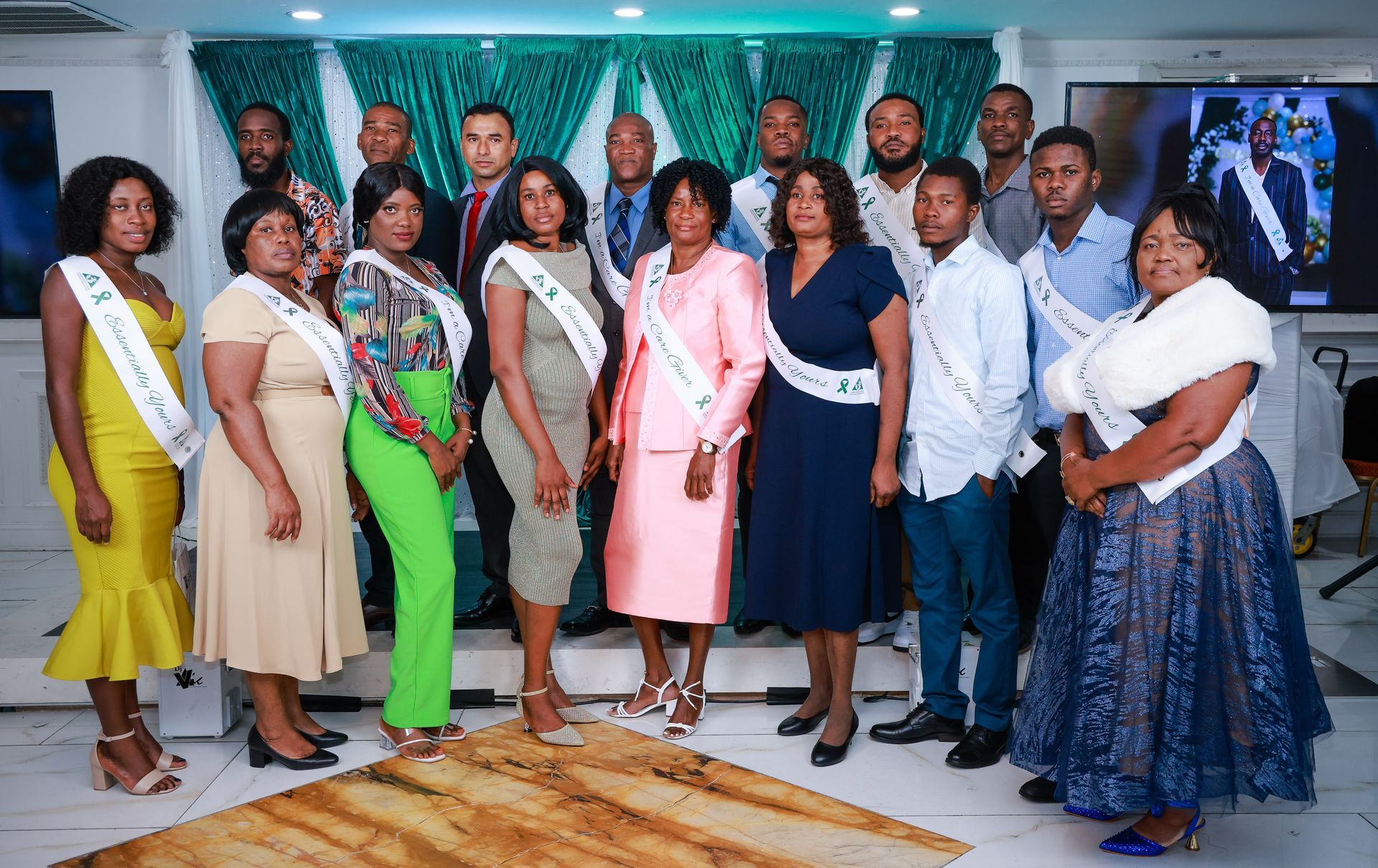 A group of people wearing sashes stand for a formal indoor portrait against a teal backdrop and decorative curtains.