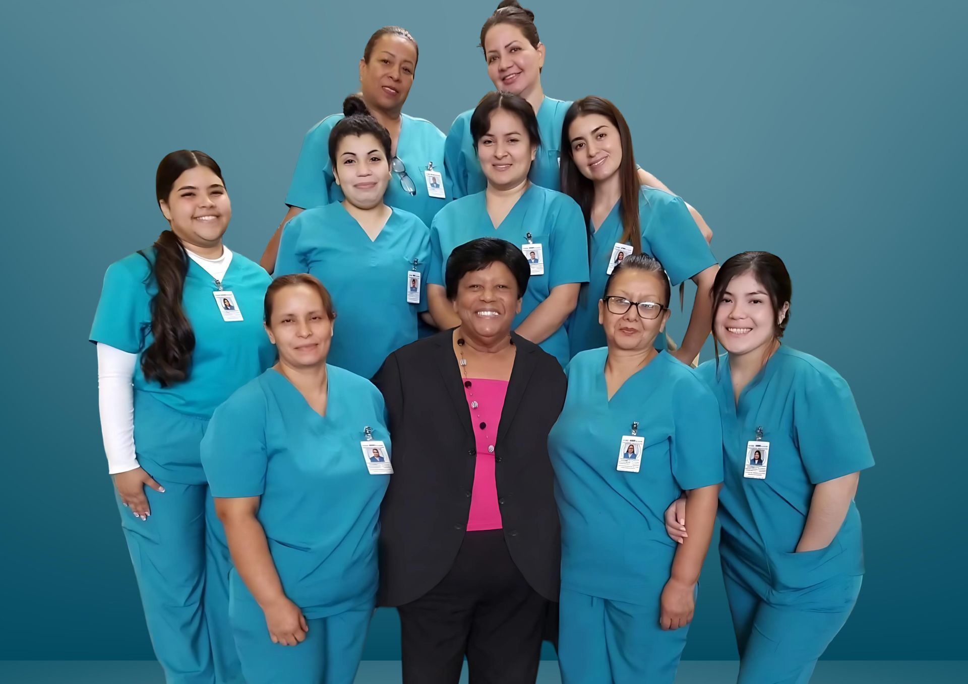 A group of ten people, nine in teal scrubs and one in a blazer and pink top, posing together against a teal background.