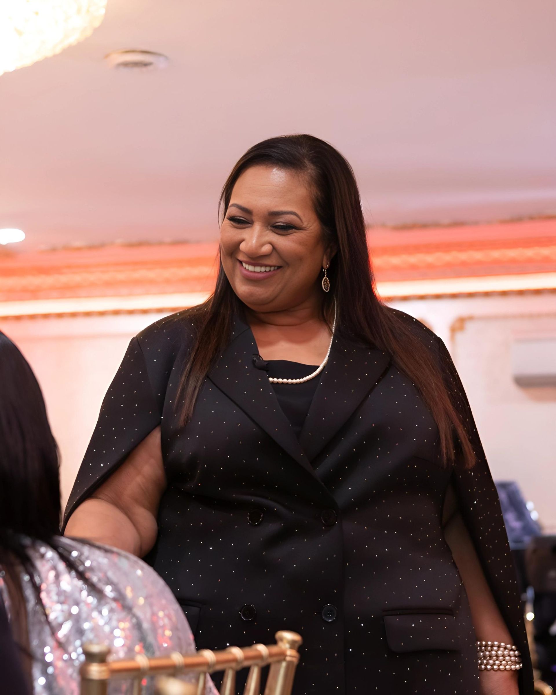 Smiling person with long hair wearing a black sparkling jacket and a necklace at an event.