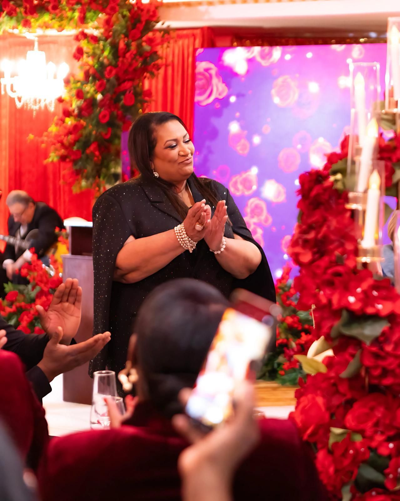 A person in a black, sparkly top applauds in a room decorated with red roses and candles while others clap in the foreground.