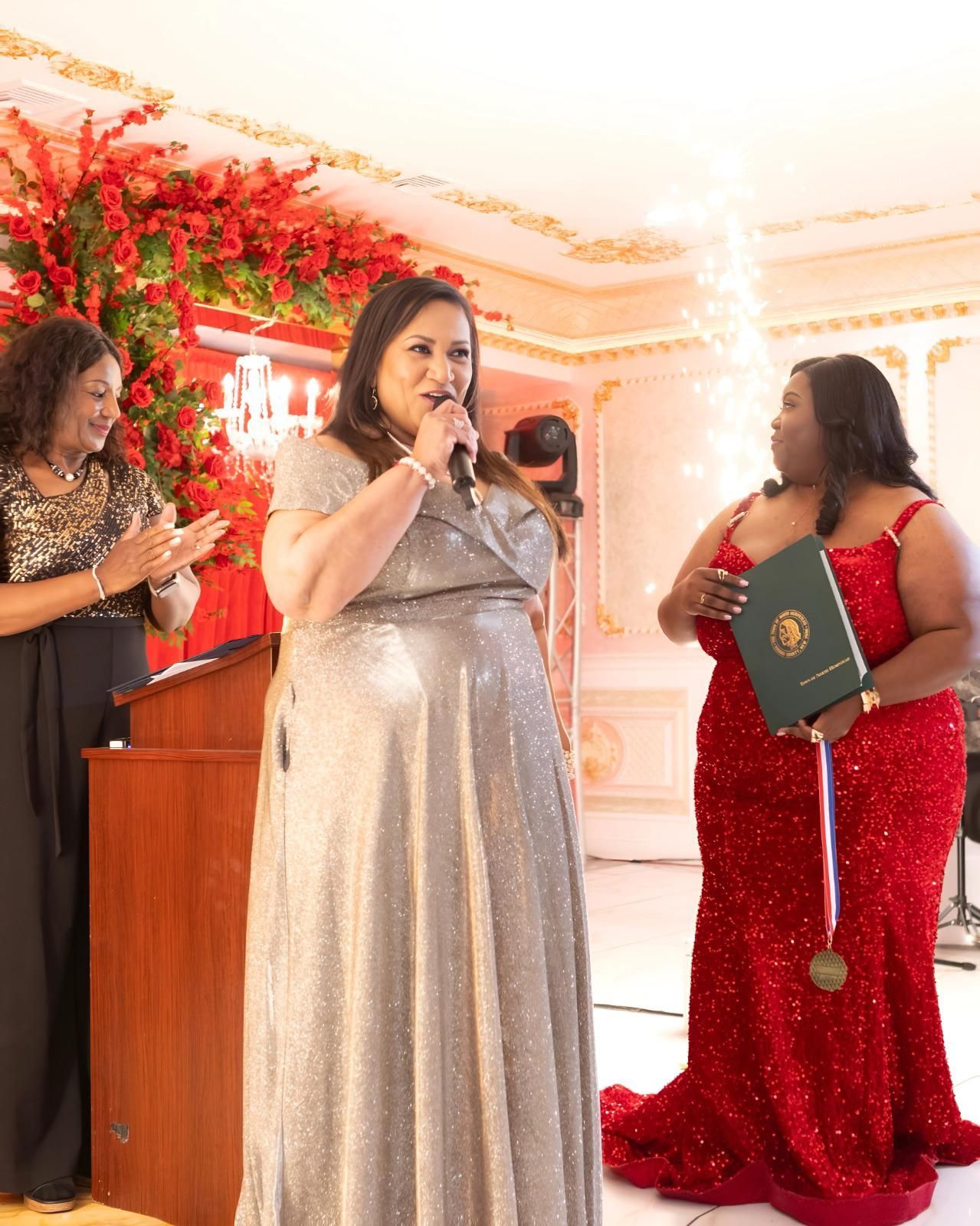 Three people in formal attire stand in a decorated hall; one speaks into a microphone while another holds a diploma case.