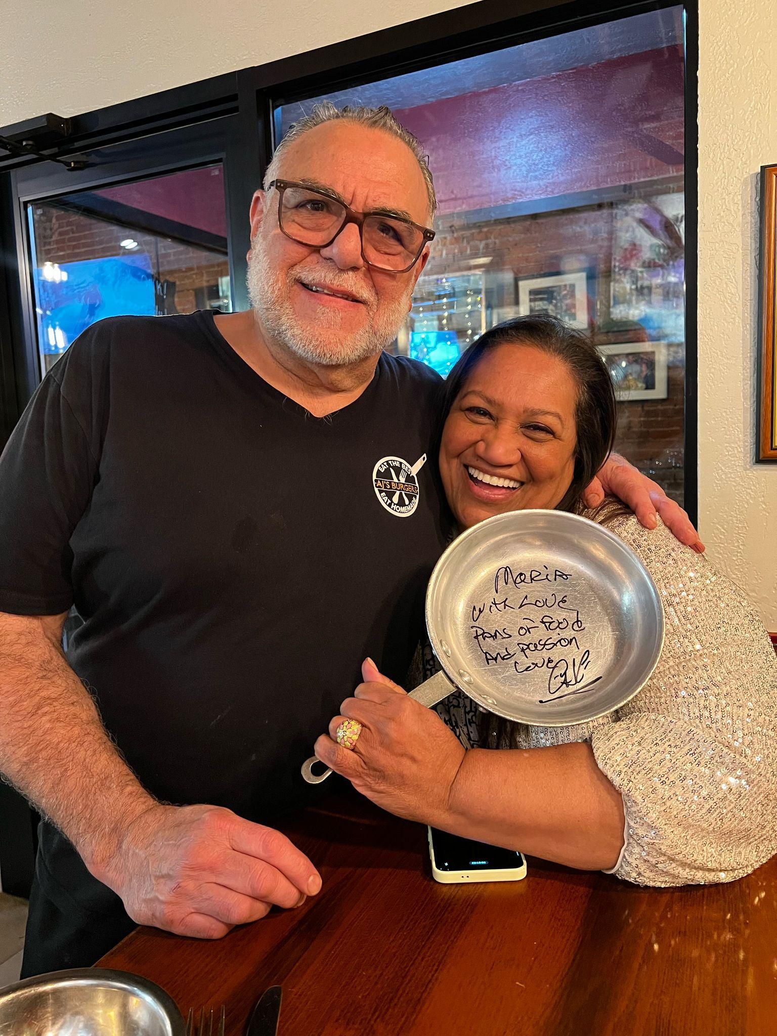 A man and woman smiling behind a counter, the woman holding a small, silver pan with handwriting inside.