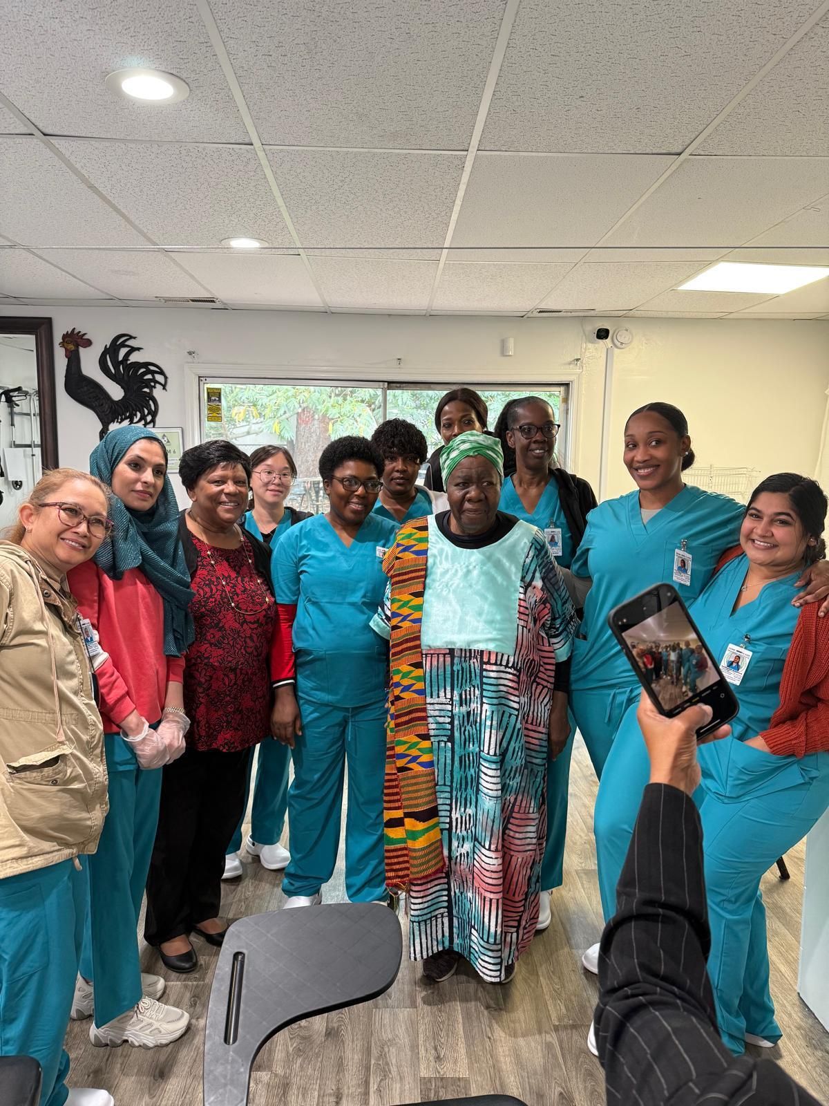 A group of people, mostly wearing teal scrubs, smiles together in a room while someone takes a photo with a smartphone.
