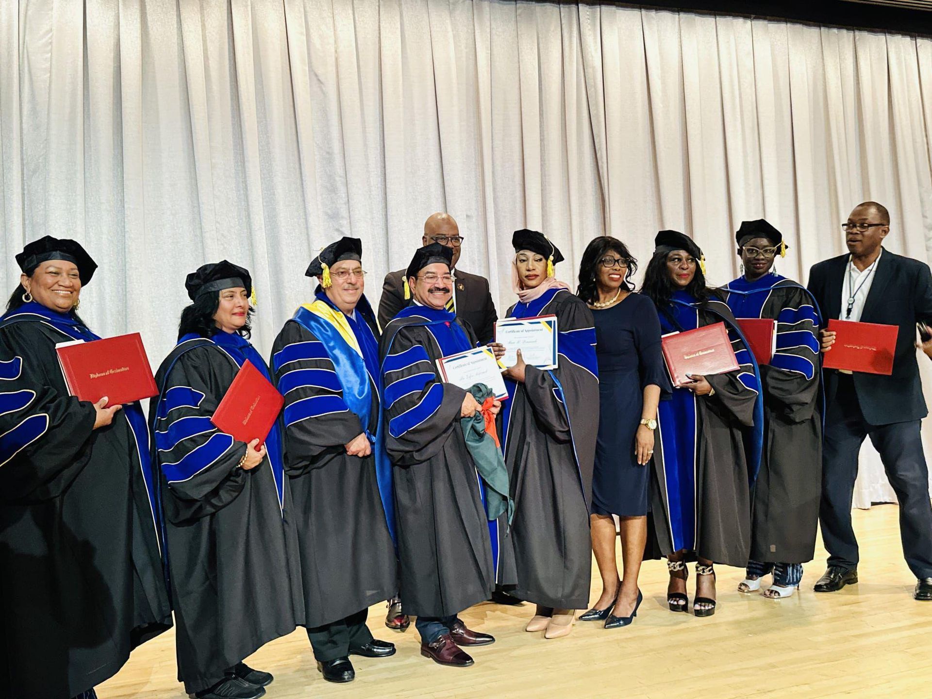 A group of individuals in academic regalia standing on a stage, some holding red folders, against a white curtain backdrop.