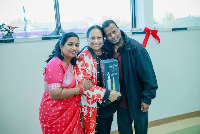 Three people stand together indoors, with the person in the middle holding a gift box.
