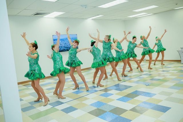 Eight dancers in matching green sequined dresses performing a choreographed routine in a brightly lit studio.