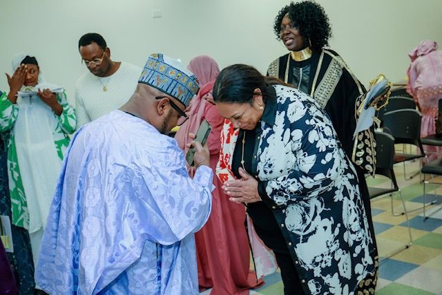 A person in a light blue patterned robe and cap bows to a woman in a black and white floral jacket in a room.