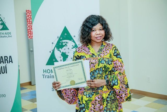 A woman holding a certificate in front of a branded Home Aide Training banner in an indoor setting.