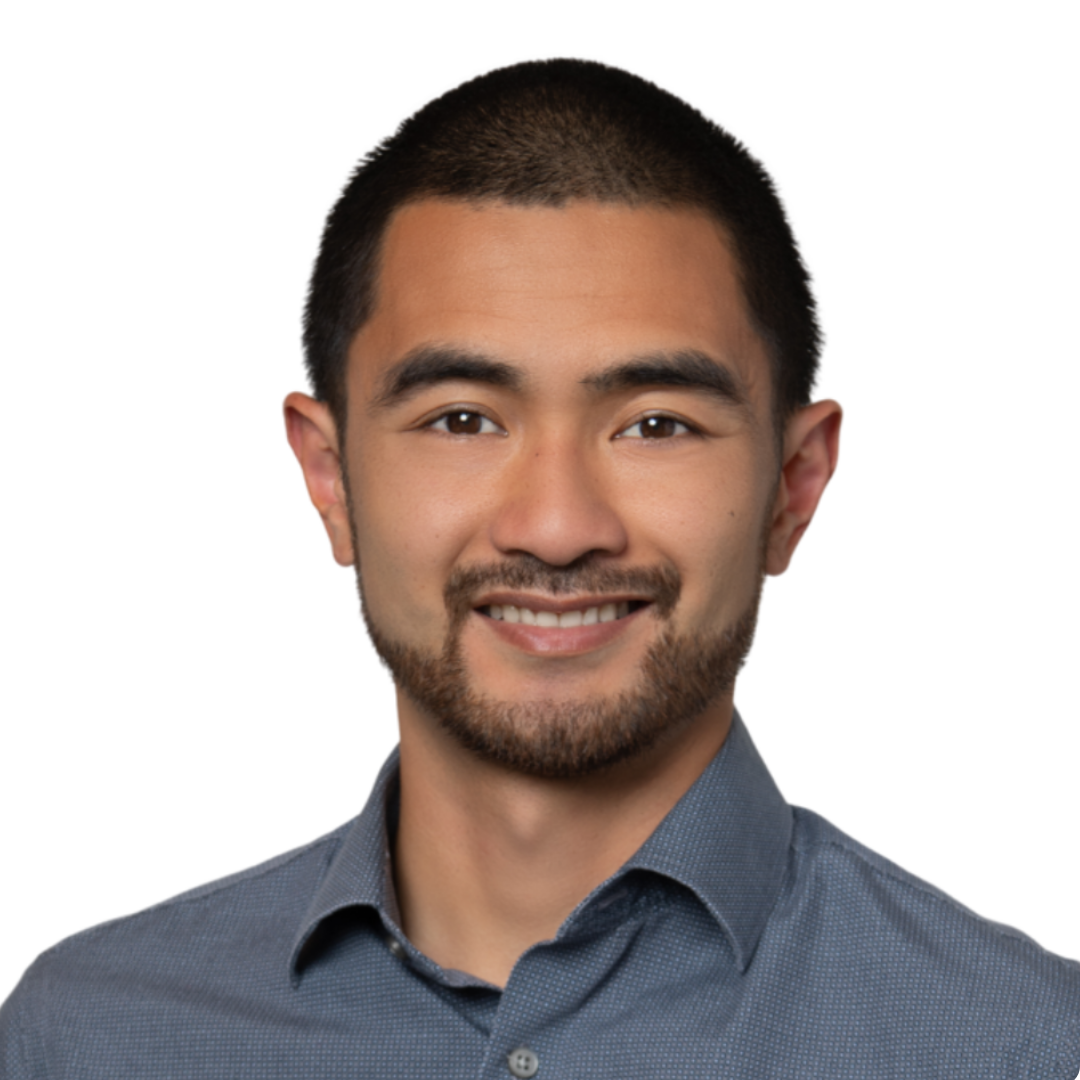 A man with short hair and a beard, wearing a blue button-down shirt, smiles against a plain light gray background.