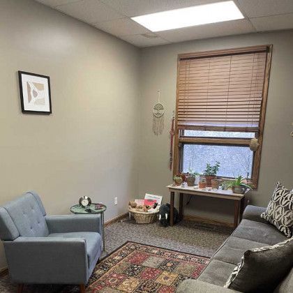 Therapy room: blue armchair, grey sofa, small table, window with plants, rug.