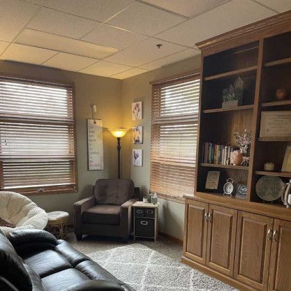 Cozy office with a brown leather couch, armchair, bookshelf, and two windows with blinds.