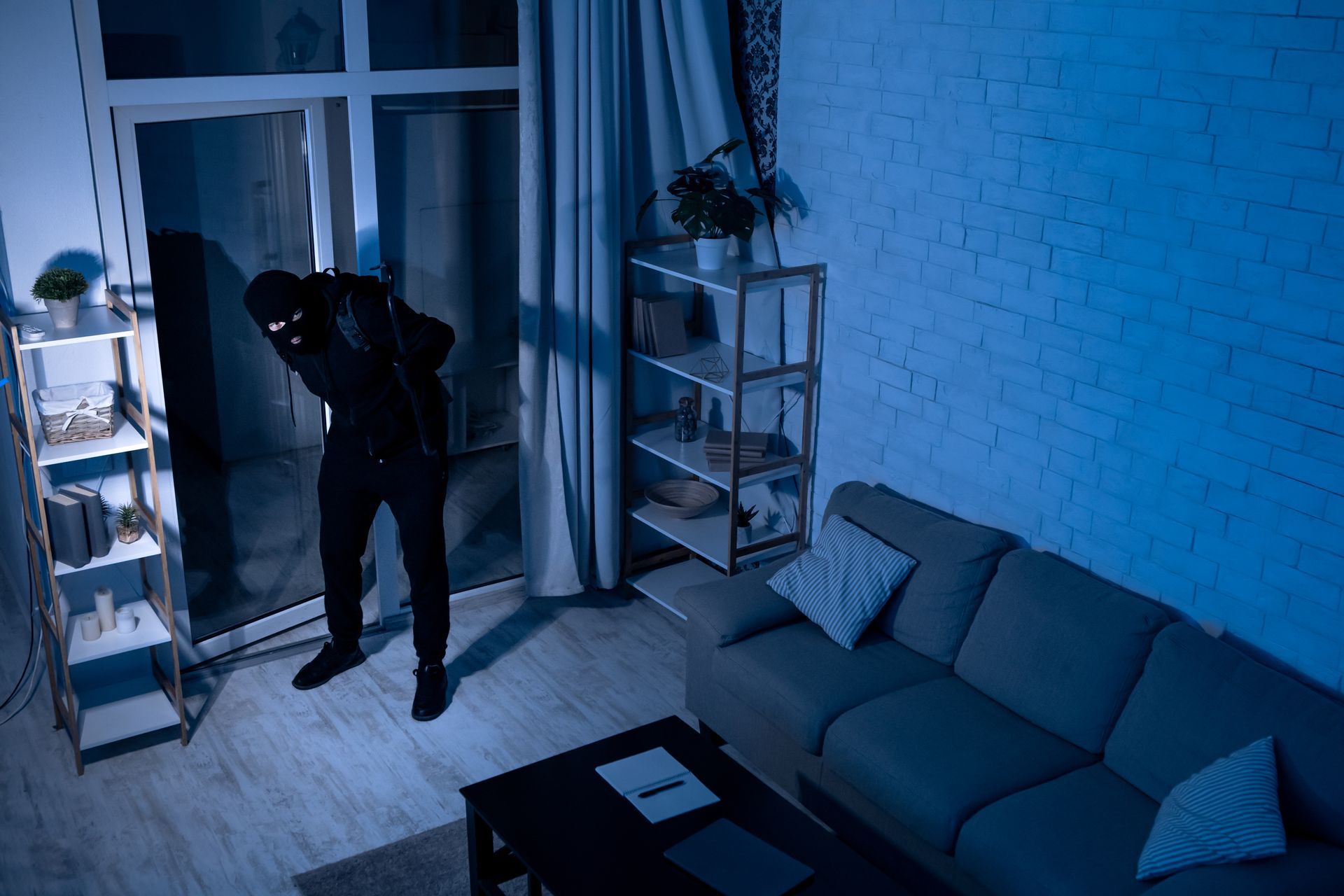 Person in dark clothing standing in a blue-lit living room near a glass door, looking down.